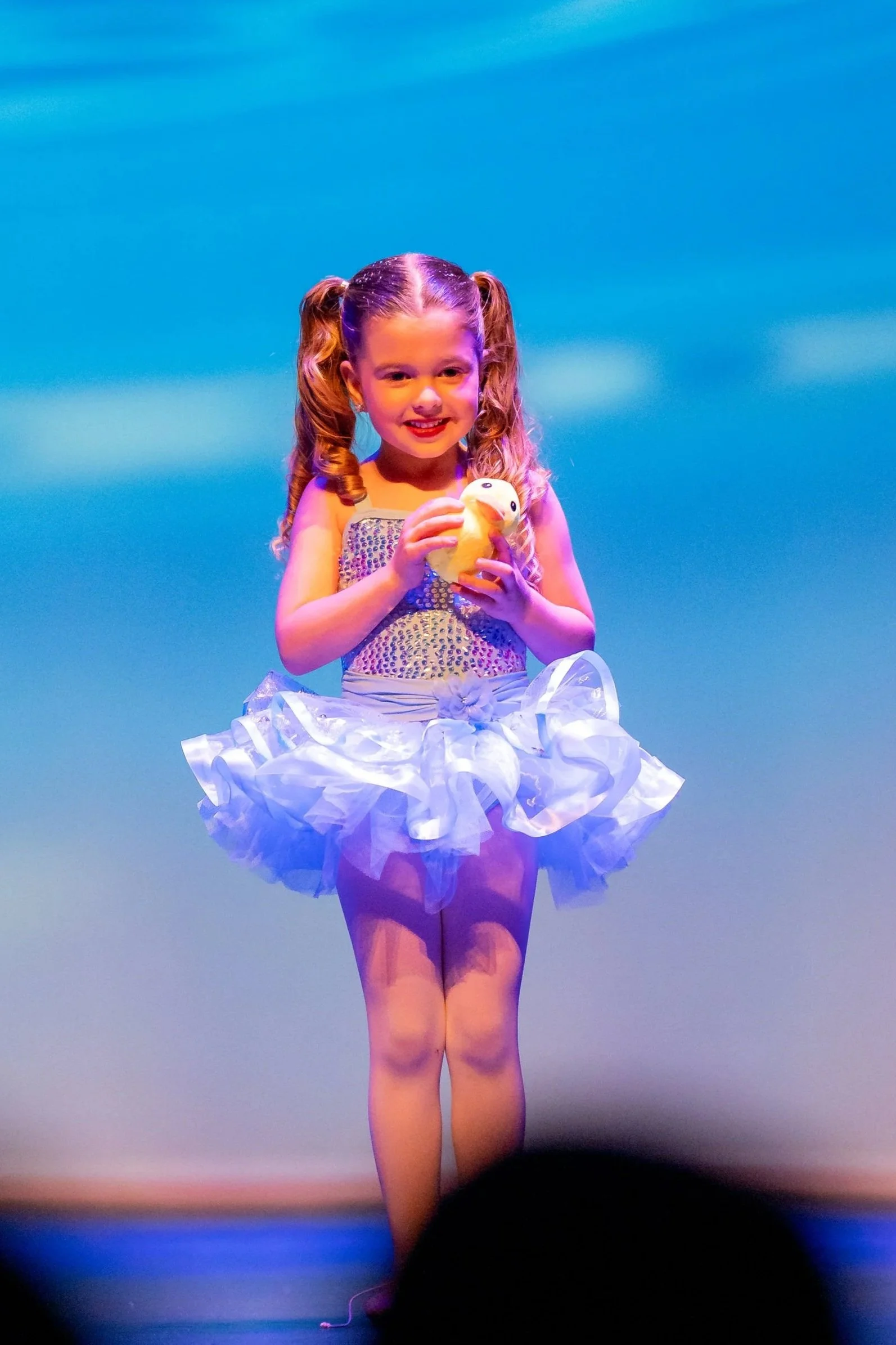 A young girl with curly hair in pigtails, smiling and holding a plush duck, standing on stage in a white tutu dress.