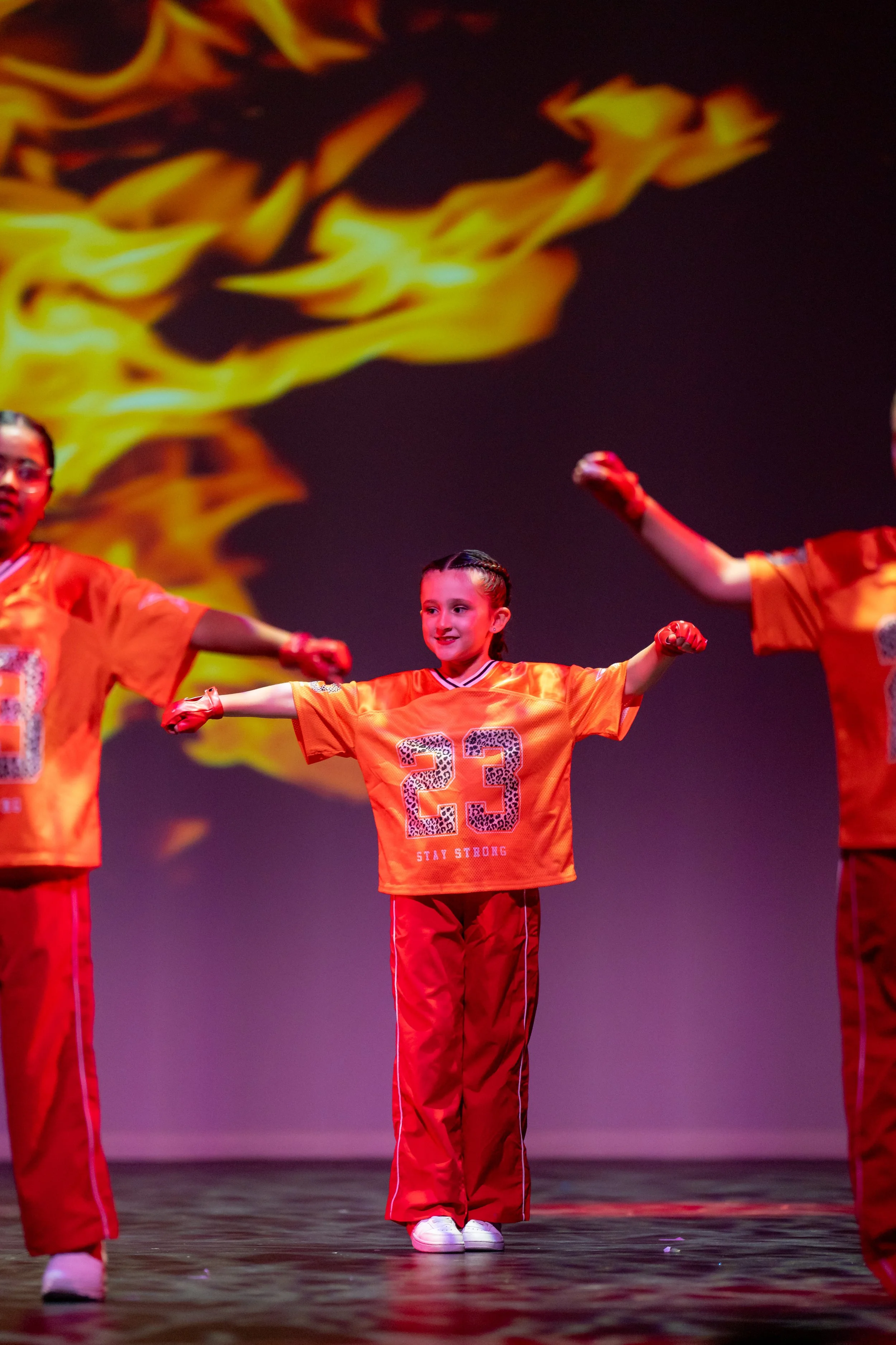 Young girl performing a dance with outstretched arms, wearing an orange sports jersey with the number 23 and the words 'Stay Strong', on stage with a fiery background projection.