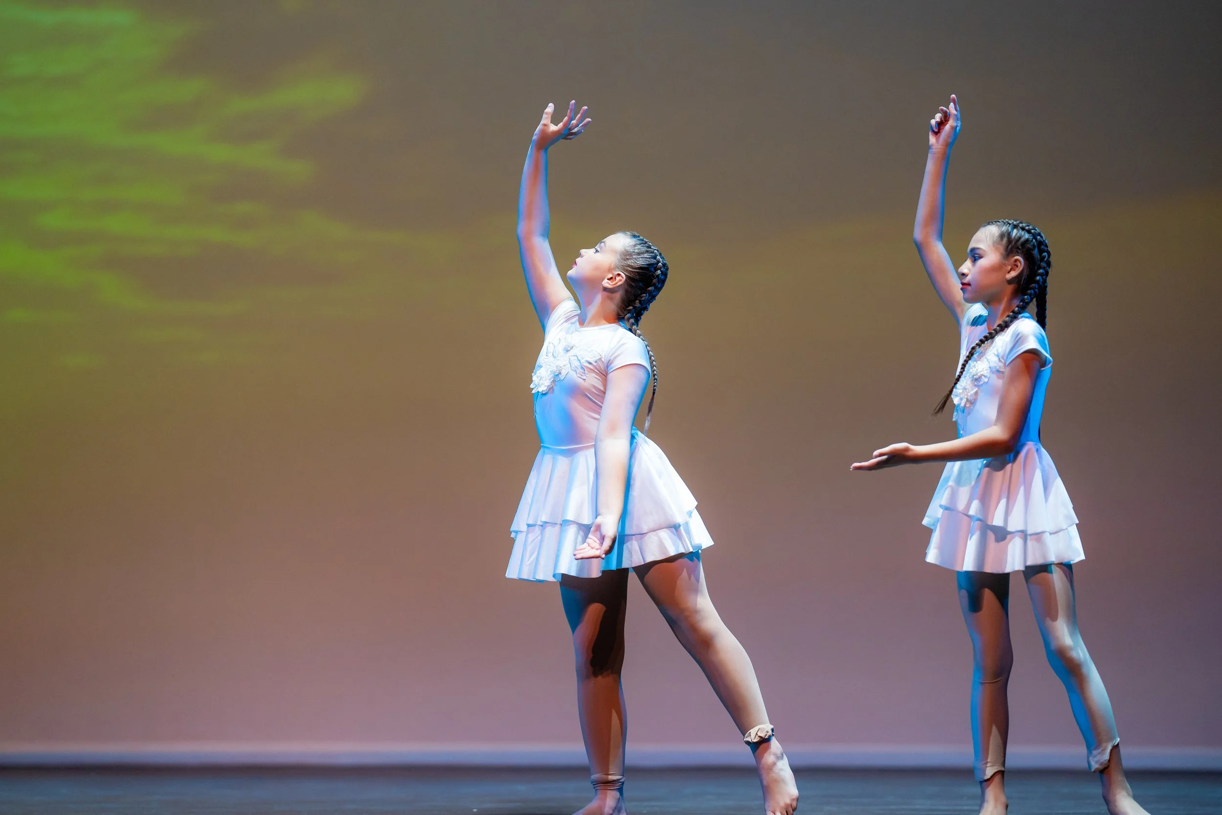 Two young girls in white ballet costumes performing a dance on stage, with their arms raised in elegant poses.