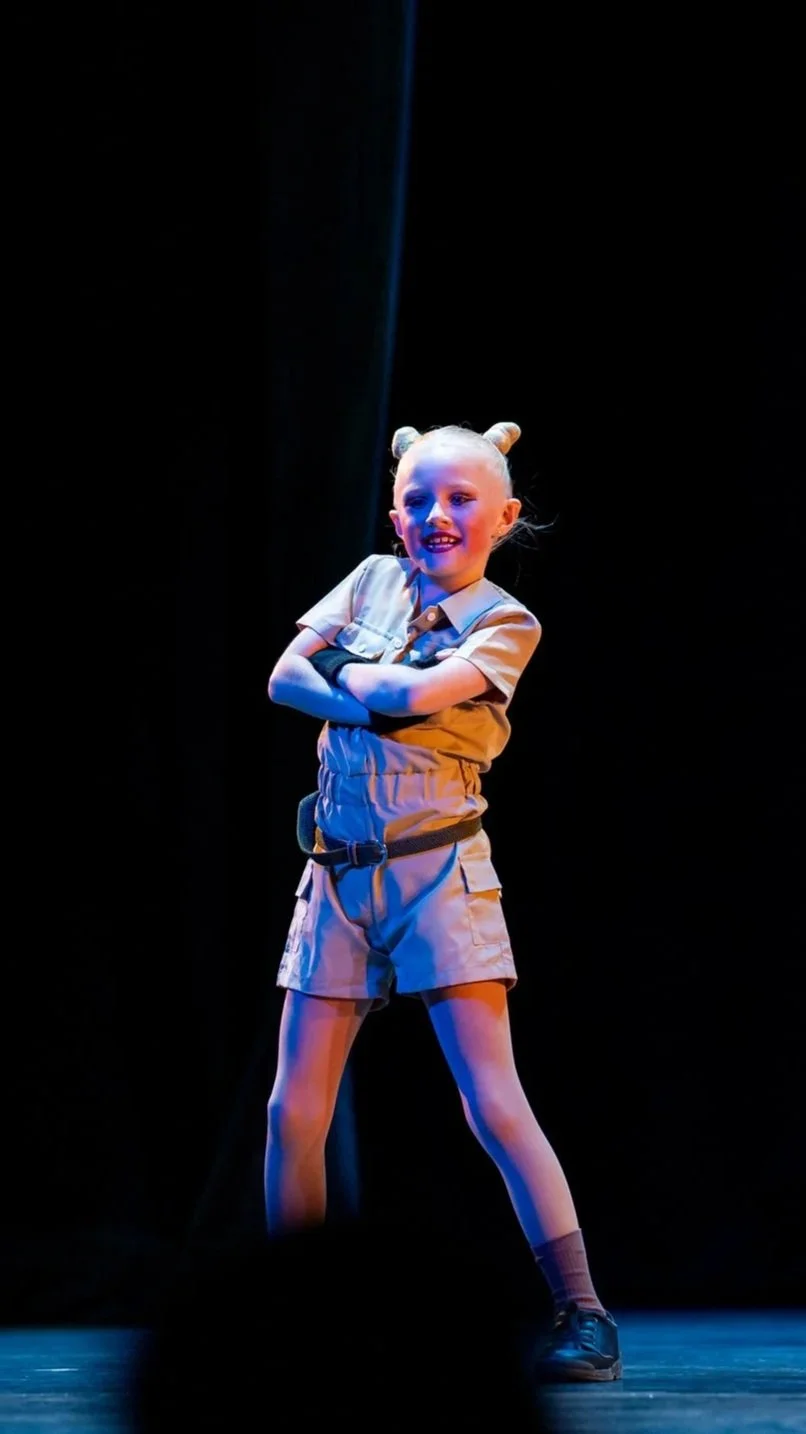 A young girl with blonde hair styled in two buns, standing on a stage with a dark background, wearing a beige short-sleeved shirt and matching shorts, with her arms crossed and smiling.