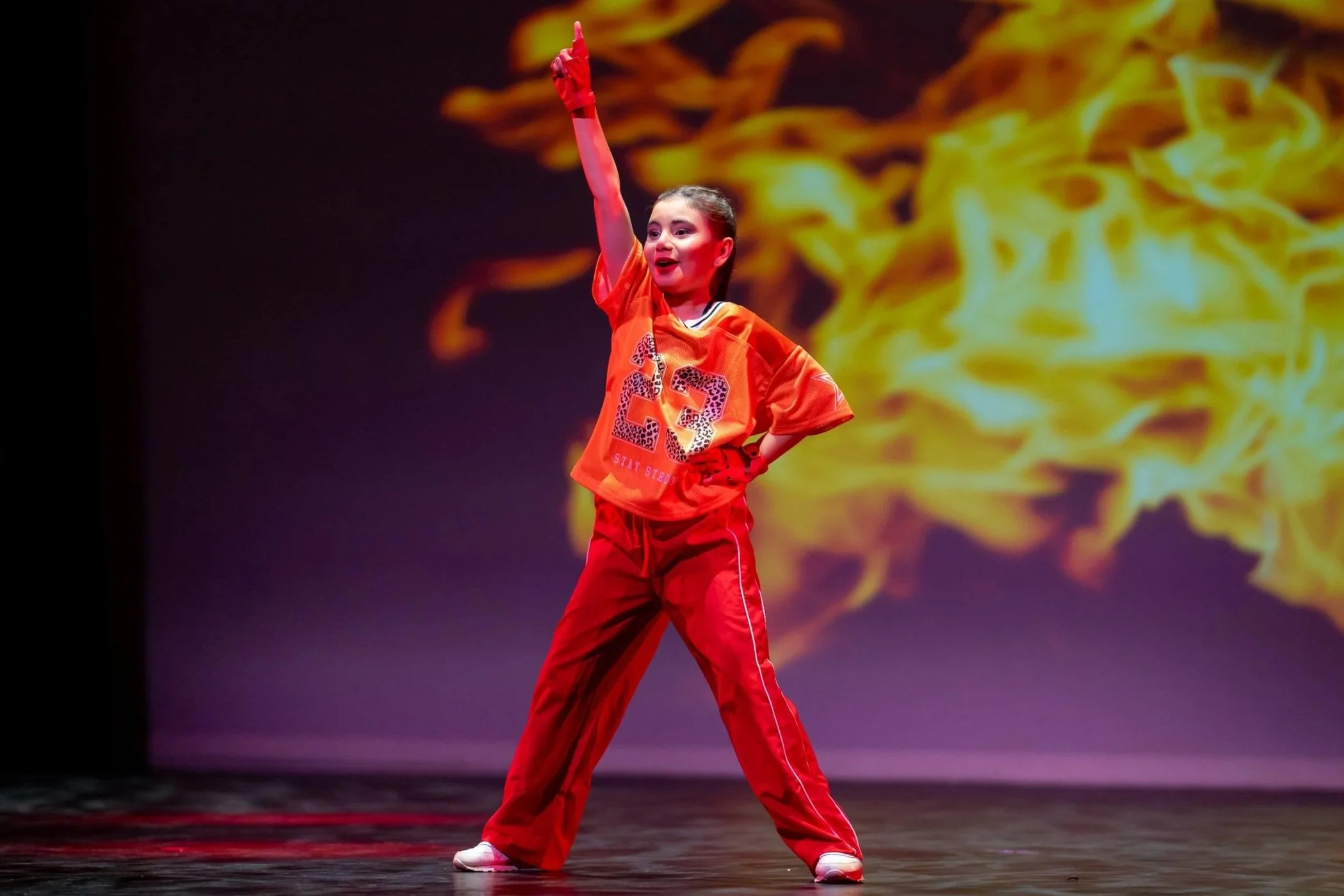 A young girl performing on stage, wearing a bright orange sports jersey with the number 23 and red pants, with a background of yellow and orange flames.