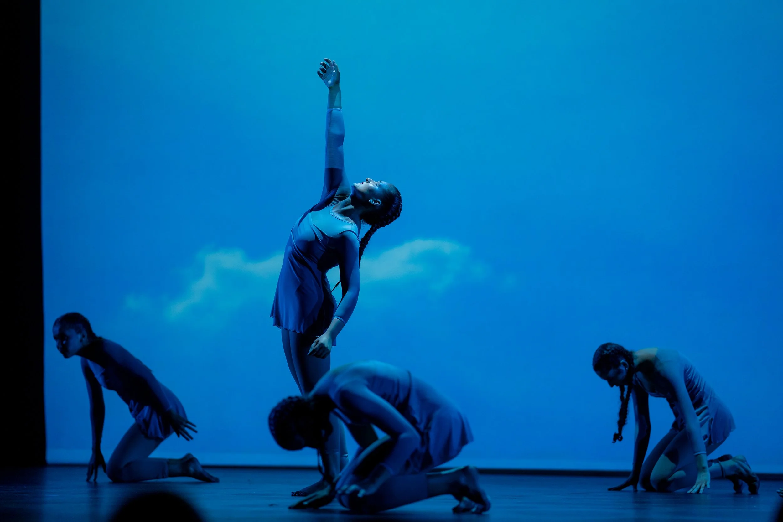 Four young ballerinas performing on stage, with one standing upright with an arm raised, and three kneeling or crouching, against a blue backdrop.
