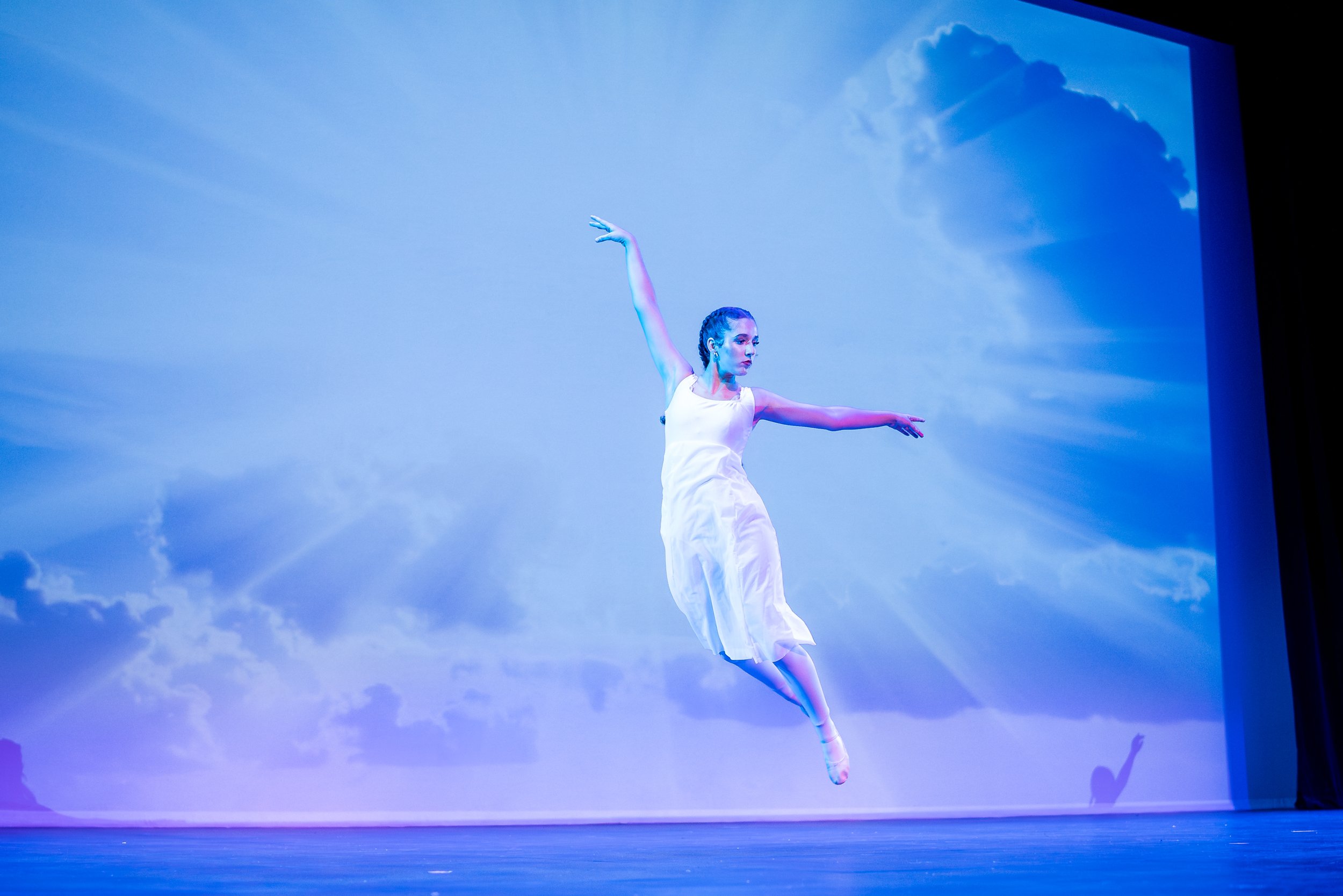 Ballet dancer in white dress performing a leap against a background of a cloudy sky projected on a screen.