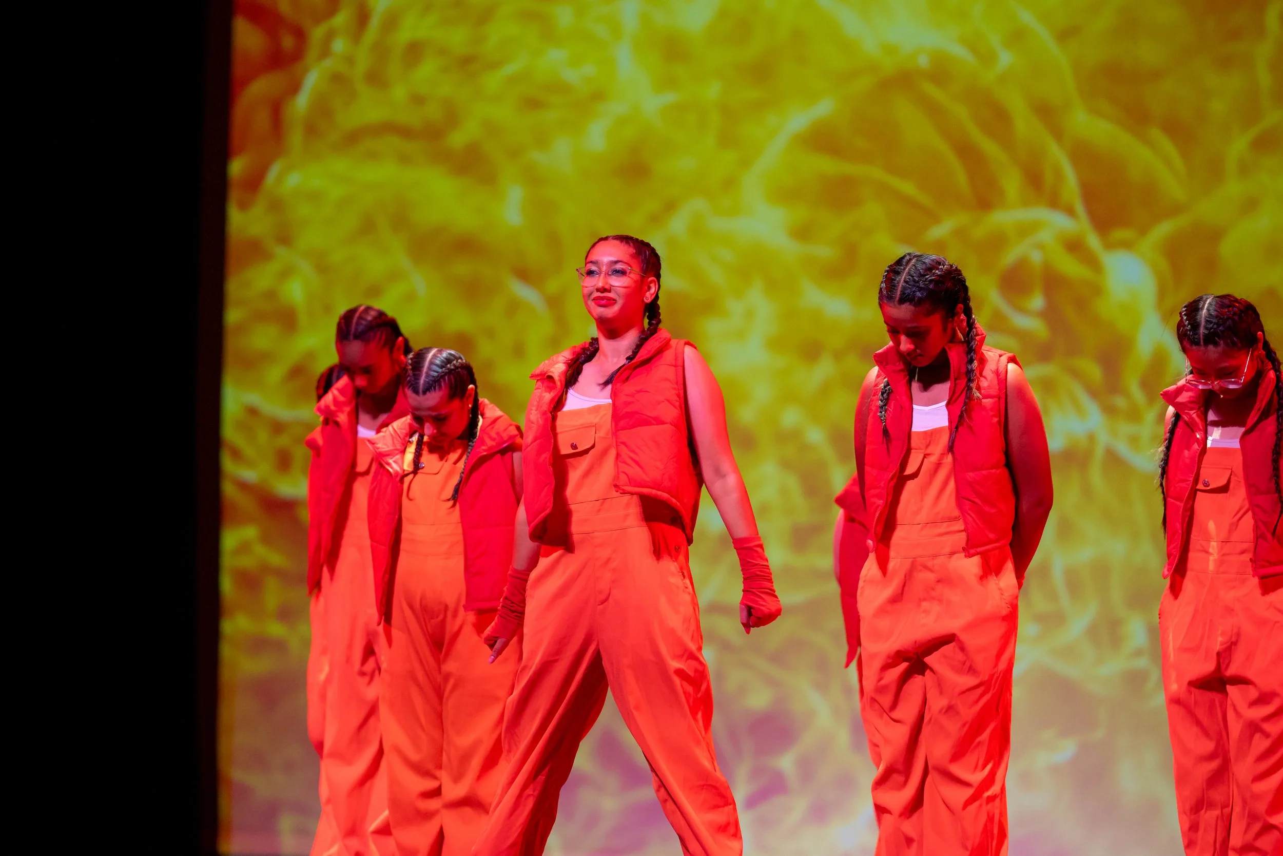 Five performers in orange jumpsuits with vests, gloves, and braids on stage with a yellow fiery background.