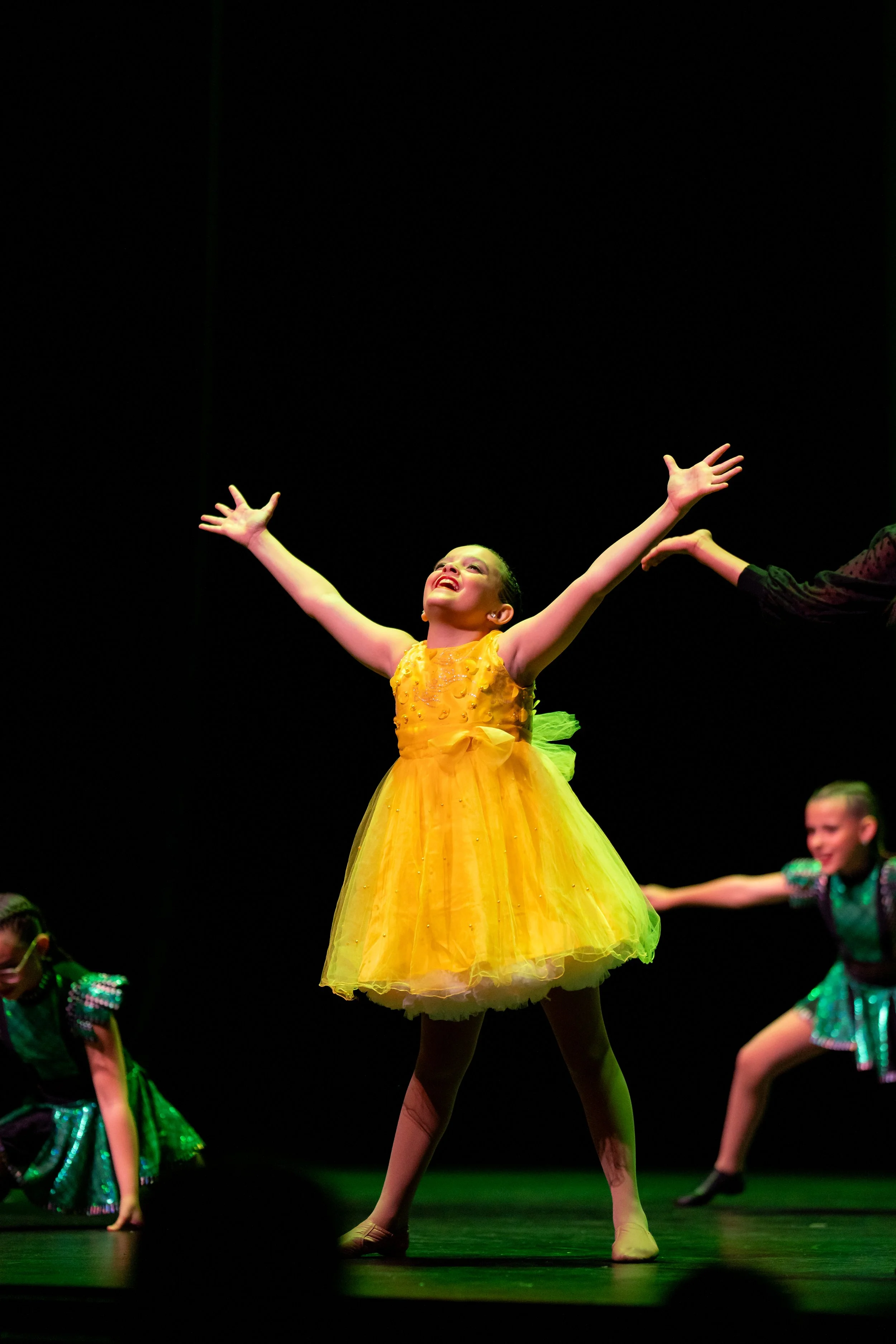 A young girl dressed in a bright yellow costume performing a dance on stage with her arms raised and a joyful expression, backed by other children in green costumes.