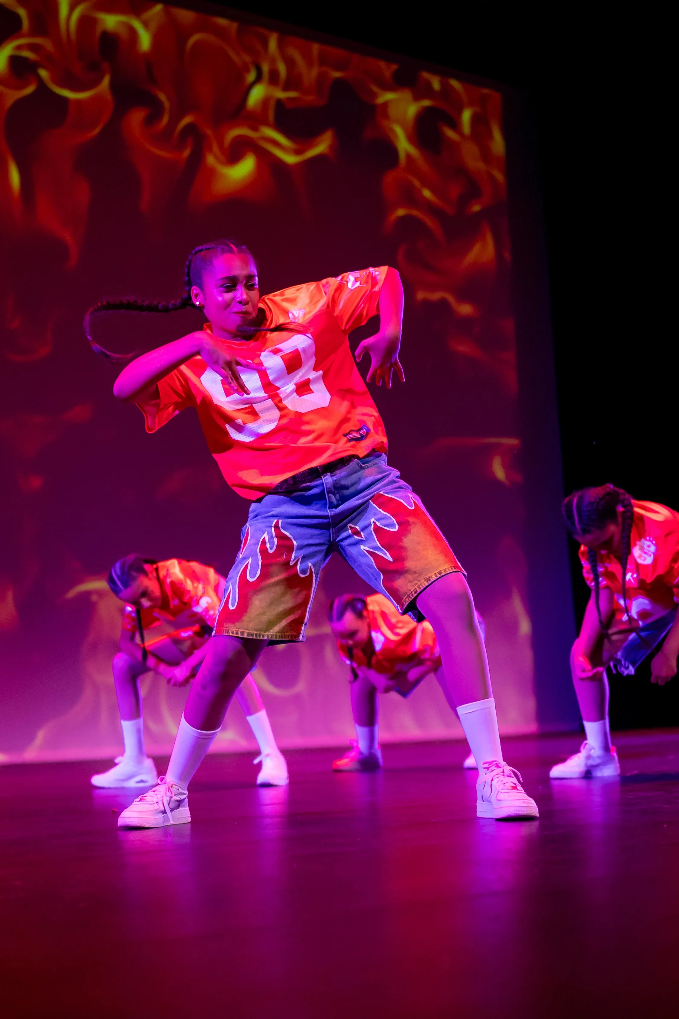 Young girl with braided hair dancing on stage with fiery background projection, wearing a red sports jersey, colorful shorts, and white sneakers.