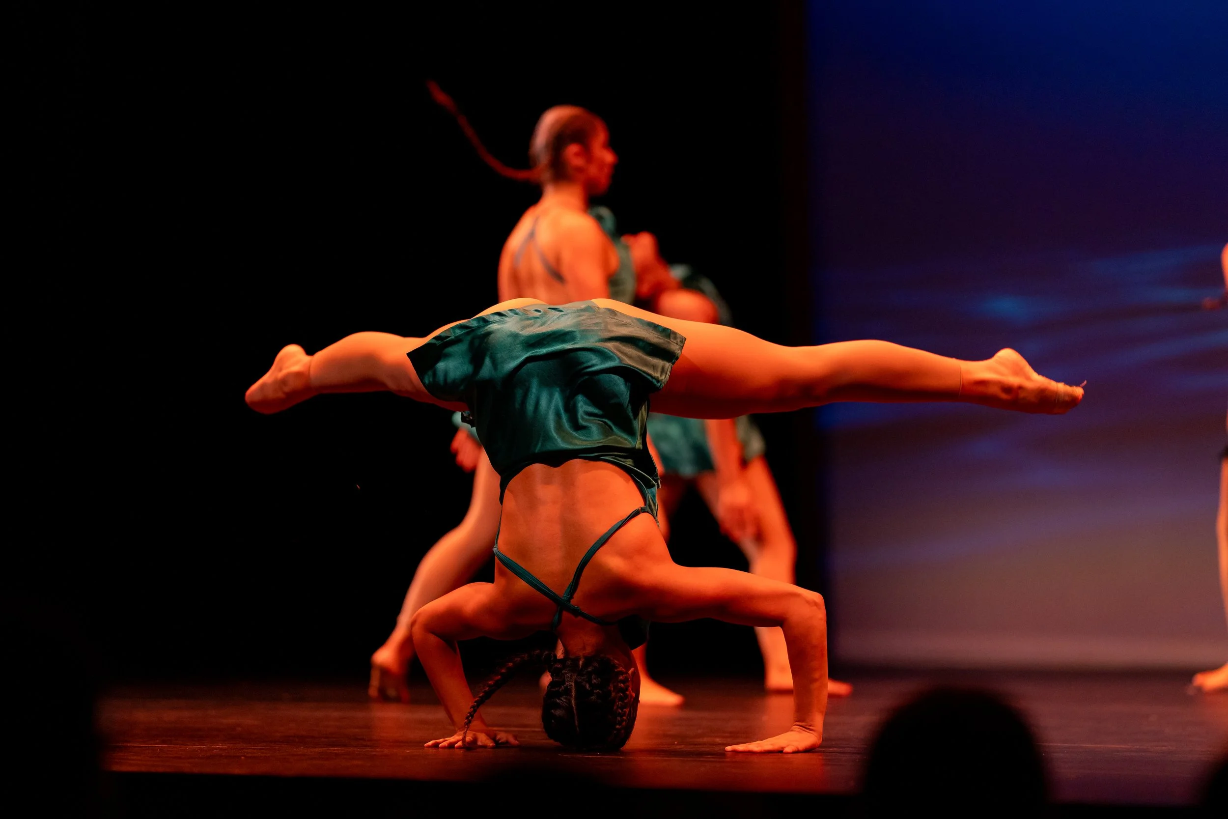 A dancer performing a headstand with legs extended in a split on a stage, wearing a green satin outfit. Other dancers are visible in the background with one blurred dancer standing with her arms crossed and a sunset scene projected on the backdrop.
