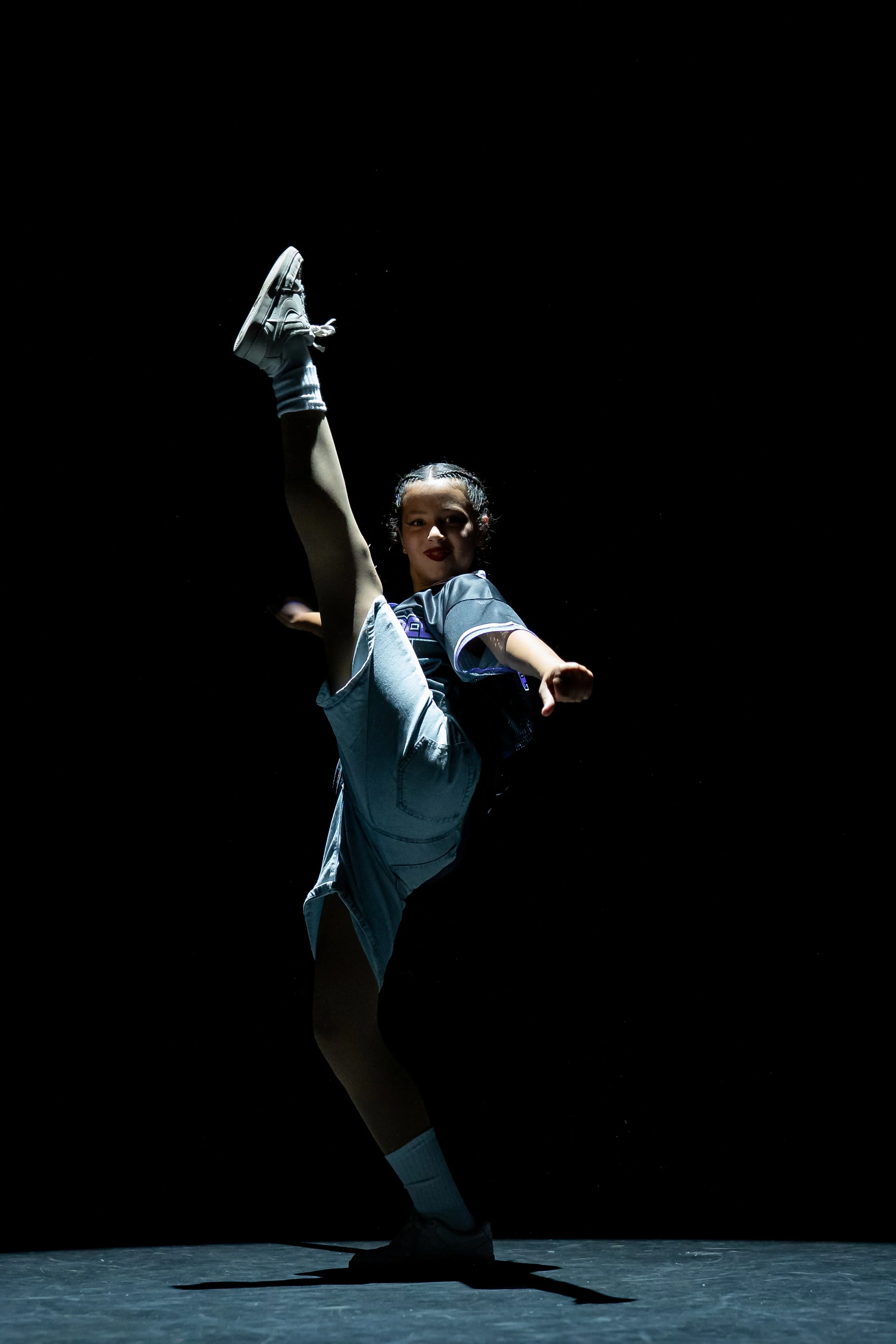 A young girl performing a high kick on stage, illuminated against a black background.