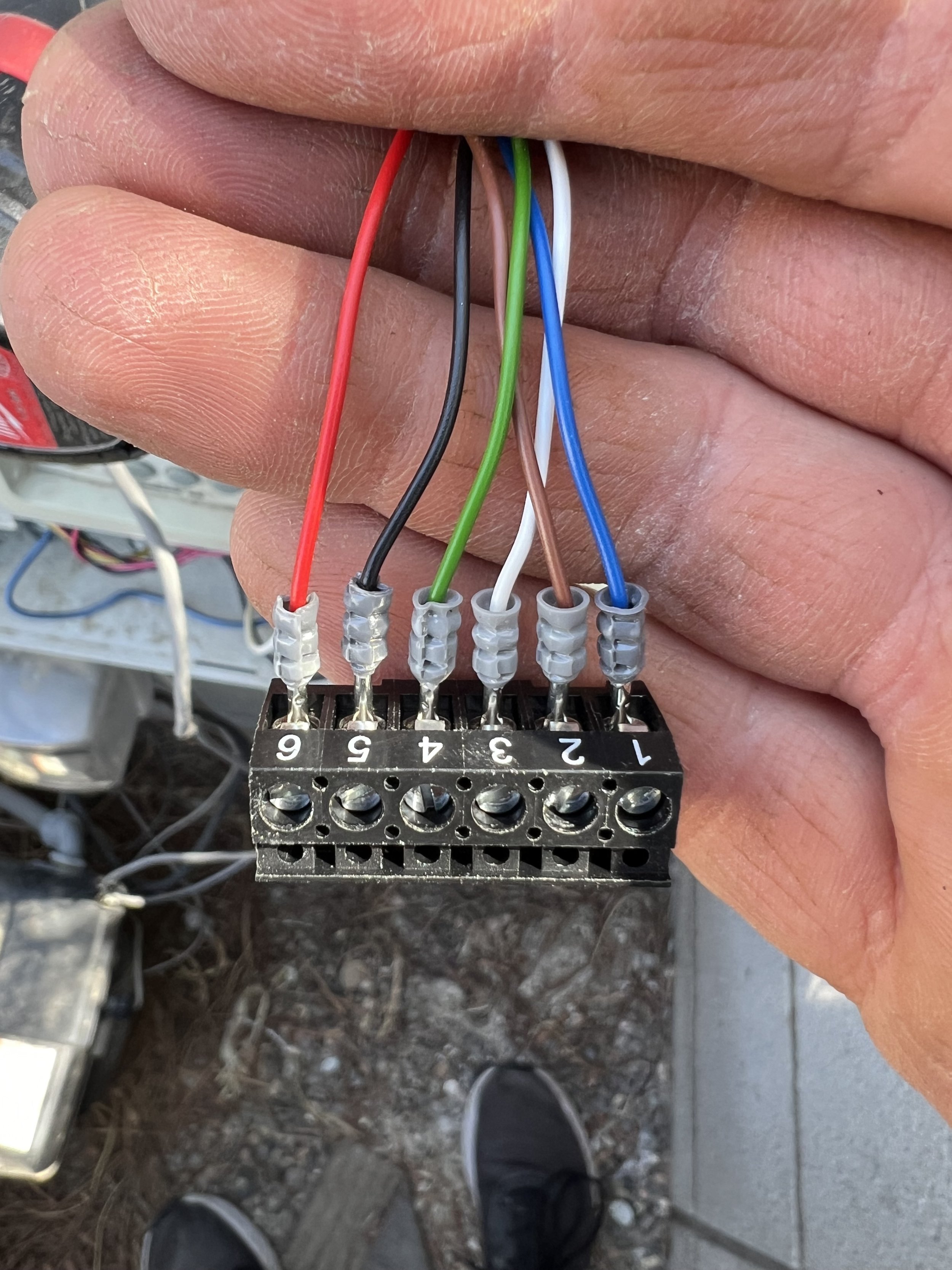 Close-up of a hand holding a terminal block connector with six colorful wires: red, black, green, white, brown, and blue, numbered 1 to 6.