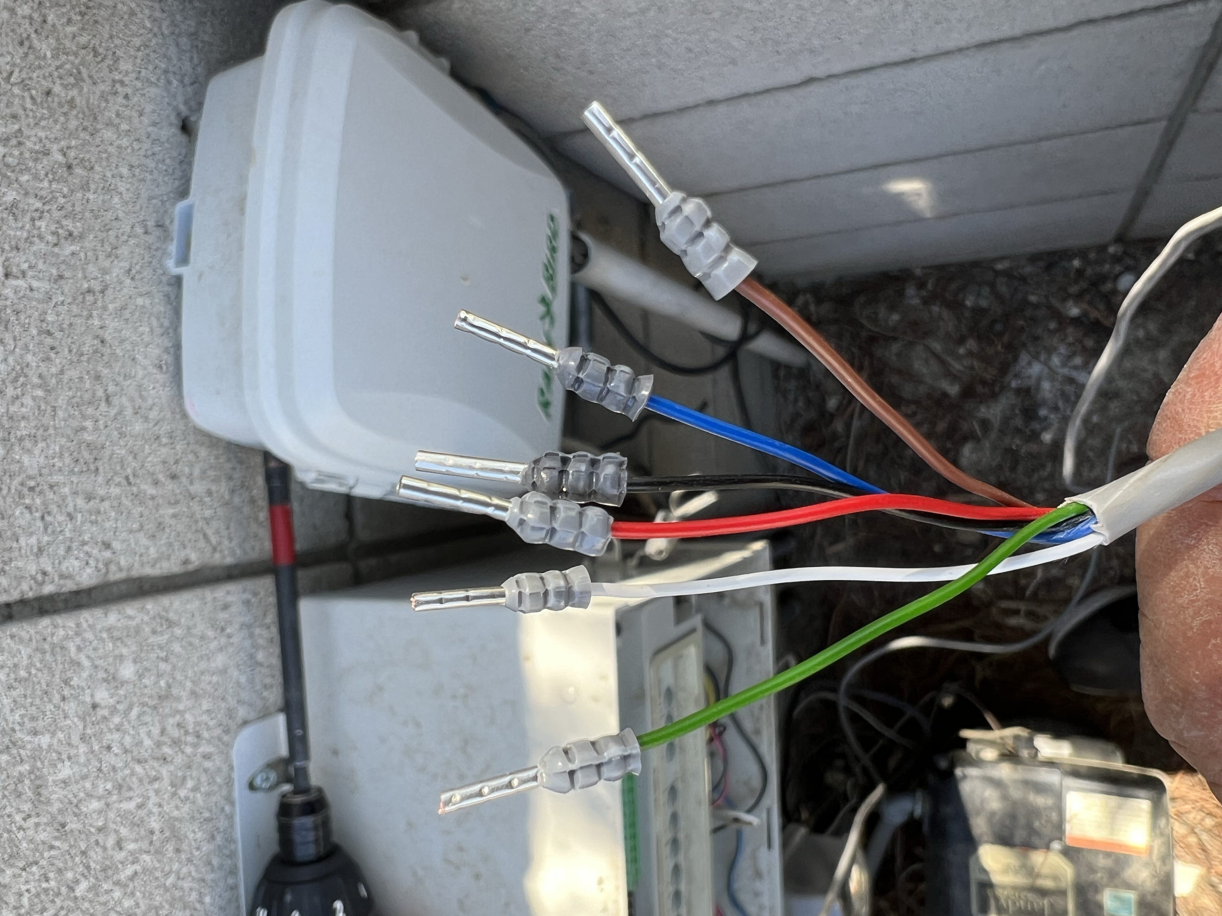 A close-up view of exposed insulated electrical wires with connectors, featuring red, green, blue, and brown wires, and a junction box in the background.