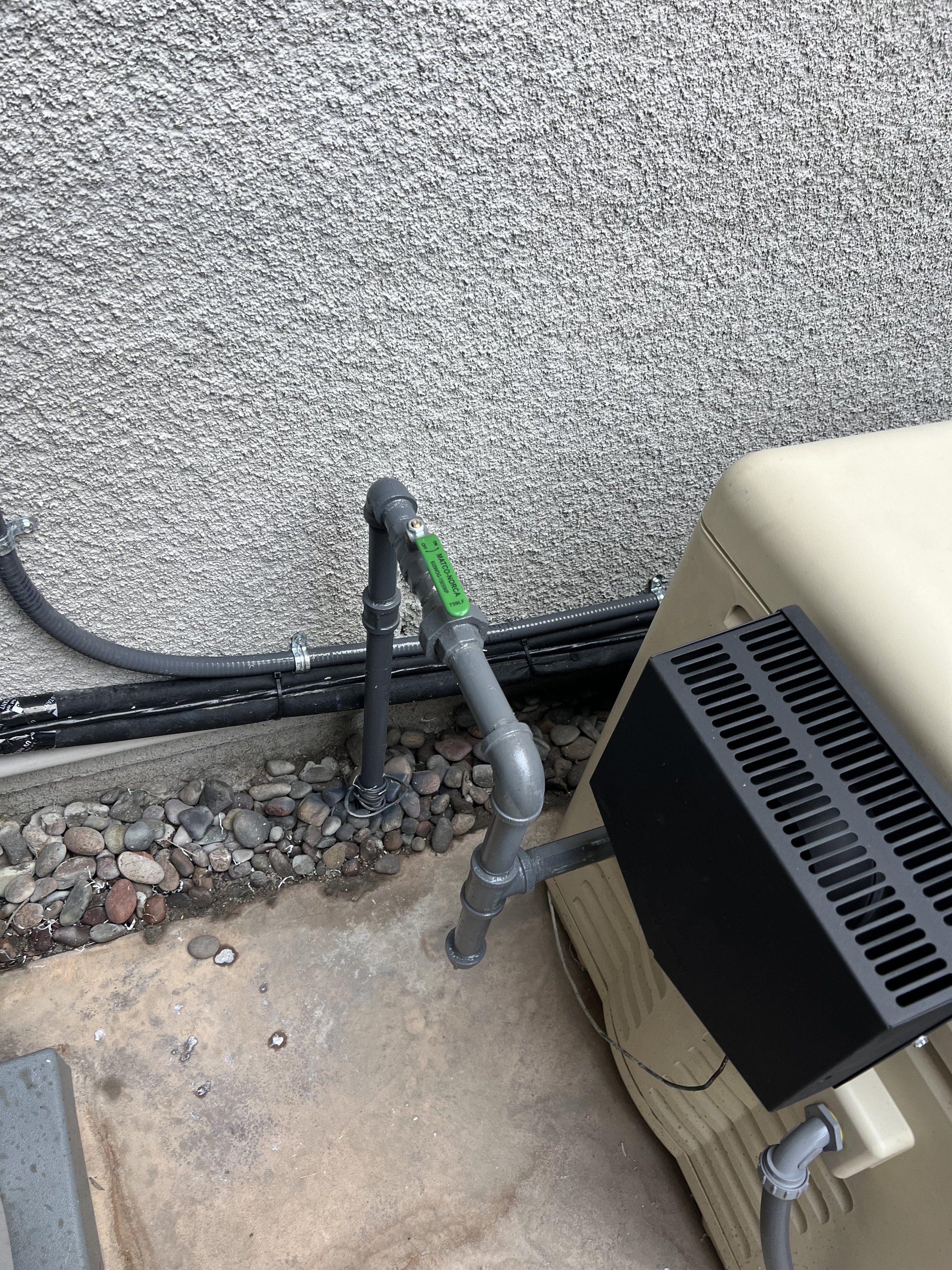 Outdoor HVAC unit with gray pipes and stones on a concrete surface next to a textured wall.