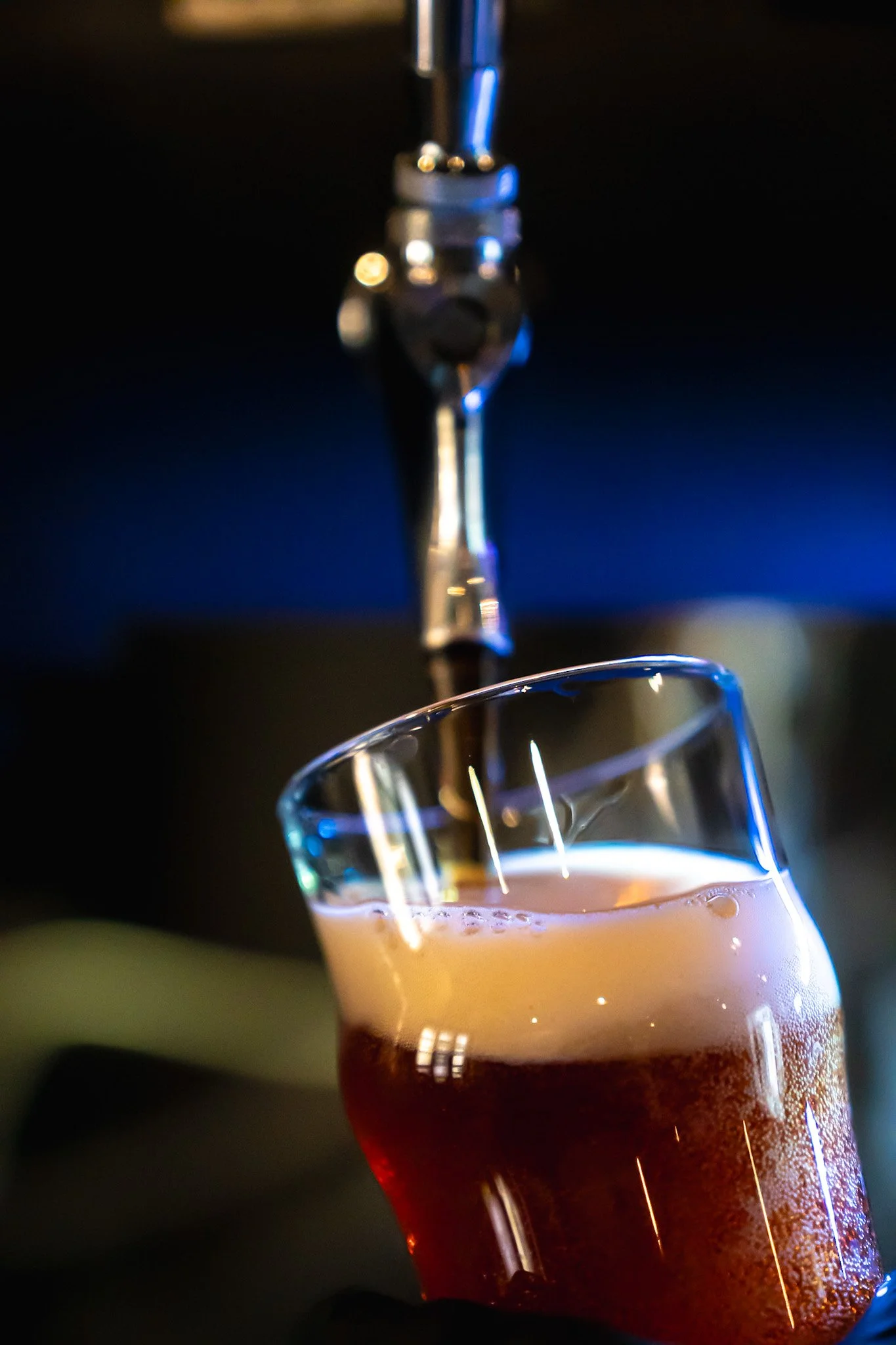 Close-up of a glass being filled with a dark amber liquid from a tap in a dimly lit setting.
