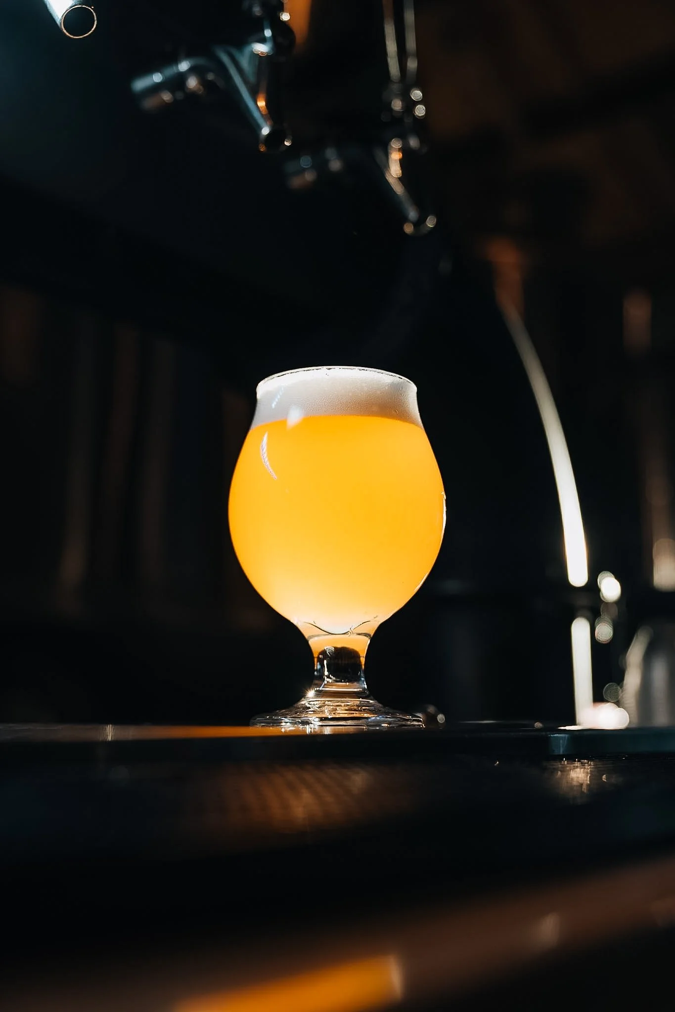 A close-up of a tulip-shaped glass filled with a hazy, yellow beer with a frothy white head, placed on a dark surface in a dimly lit setting.