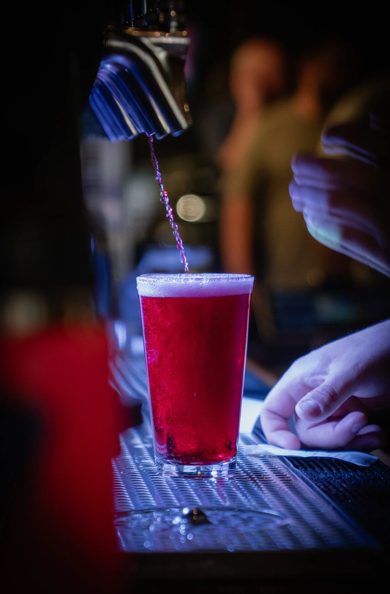 A bartender is pouring a red liquid from a tap into a glass at a bar, with a focus on the glass and hand, blurred background.