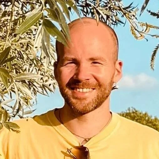 A young man with a shaved head, beard, and mustache looks at the camera with a slight smile outdoors on a sunny day.
