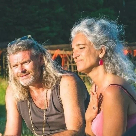 A woman and a man sitting outdoors in a green environment, smiling and enjoying each other's company.