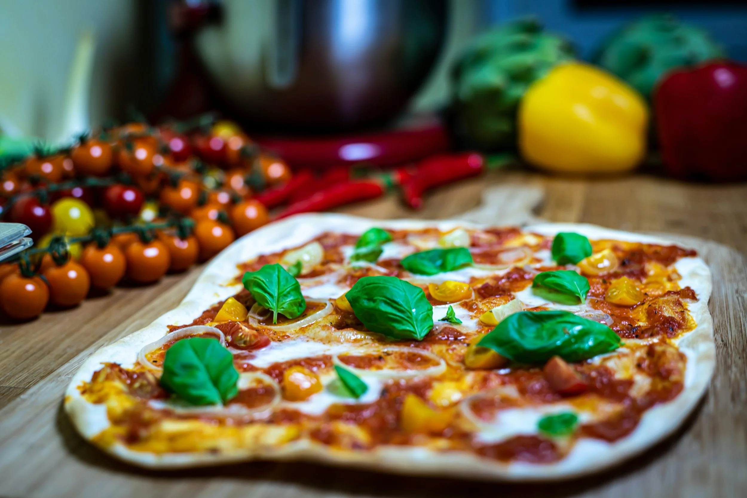 Frische Pizza mit Basilikum, Tomaten und Zwiebeln auf Holzbrett, im Hintergrund bunte Paprika, Tomaten und Chilis.
