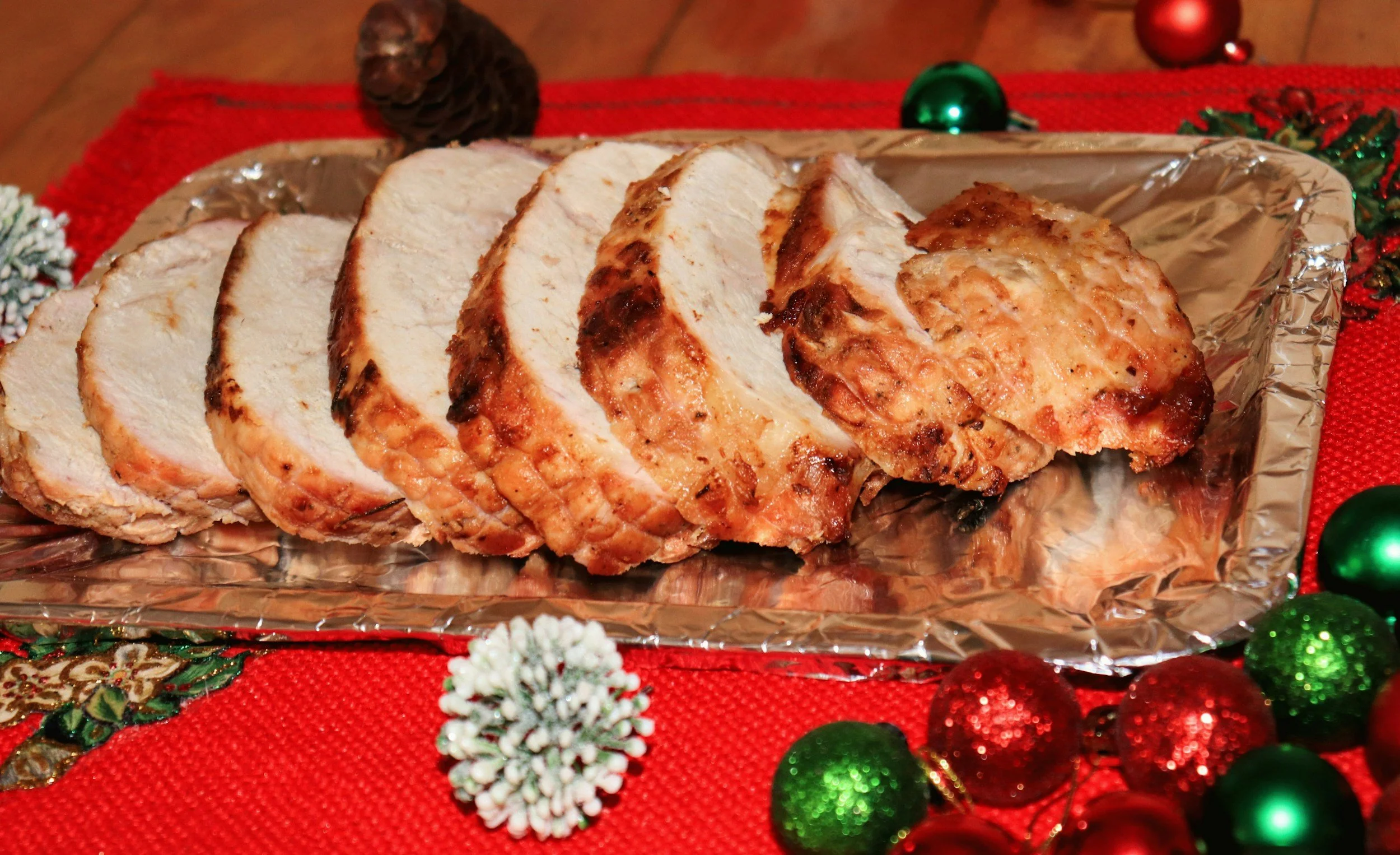 Auf einem silbernen Tablett liegt in Scheiben geschnittener Braten, wahrscheinlich Putenbrust, auf einem roten festlichen Tuch, umgeben von Weihnachtsdekorationen wie Kugeln und Schneeflocken.