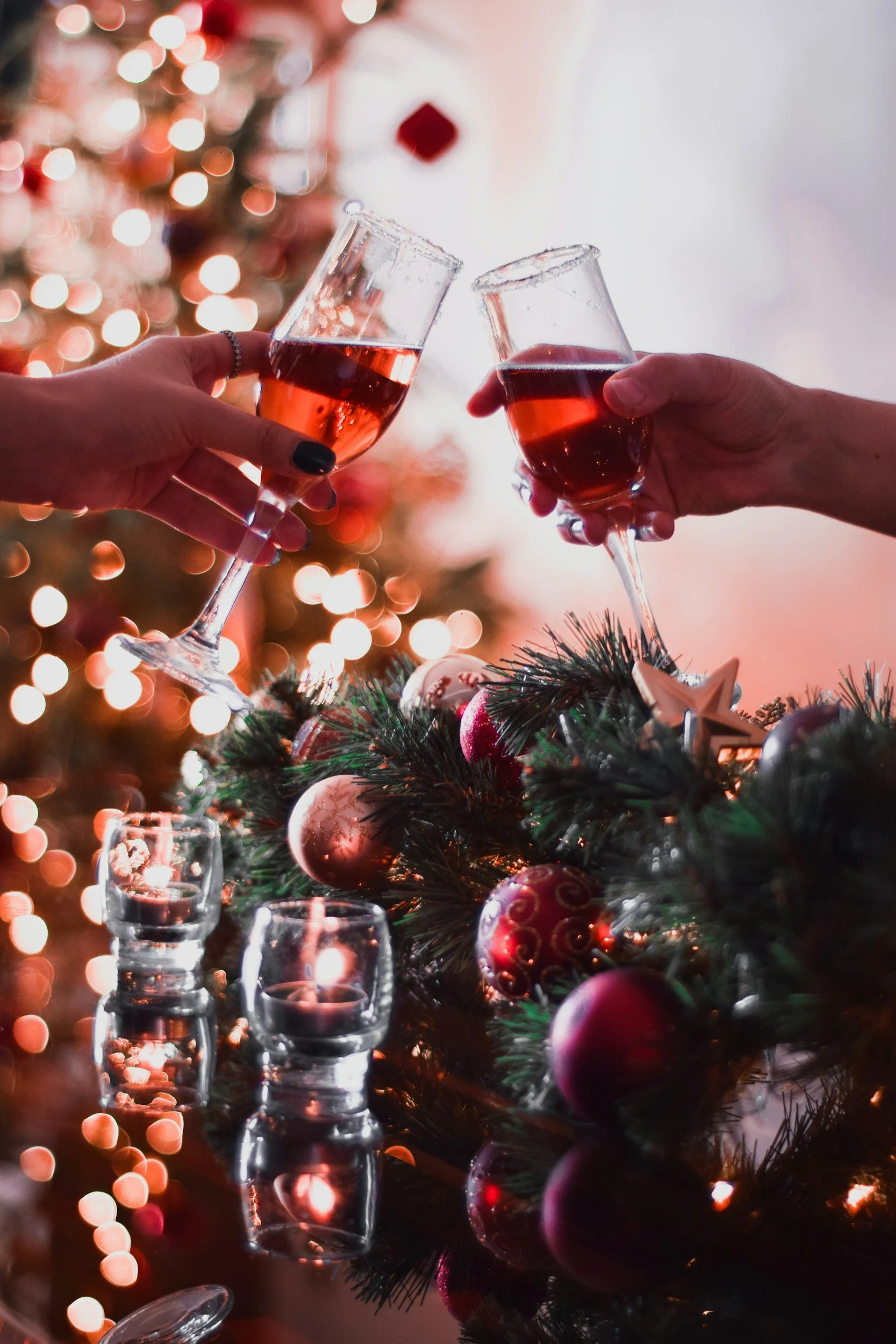 Zwei Personen stoßen mit Weingläsern mit rotem Wein an, im Hintergrund ein geschmückter Weihnachtsbaum mit Lichtern, und im Vordergrund eine Weihnachtsdekoration mit Kerzen, Ornamenten und Tannenzweigen.