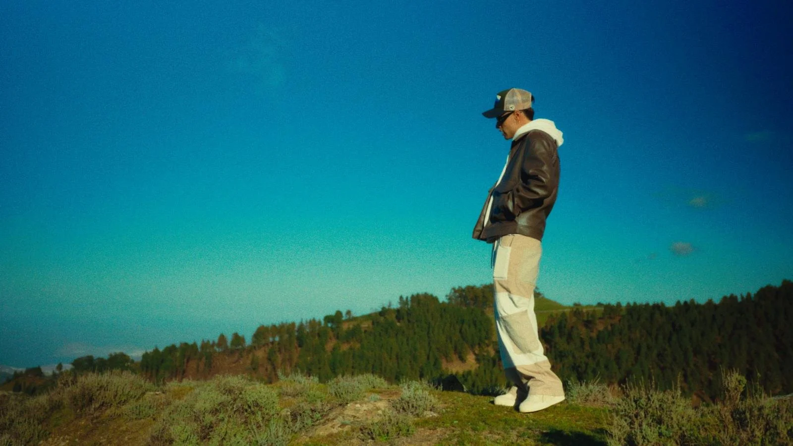 Joven vestido con chaqueta marrón, pantalones claros y gorra, de pie en un campo, mirando hacia abajo, con un fondo de árboles y un cielo azul.