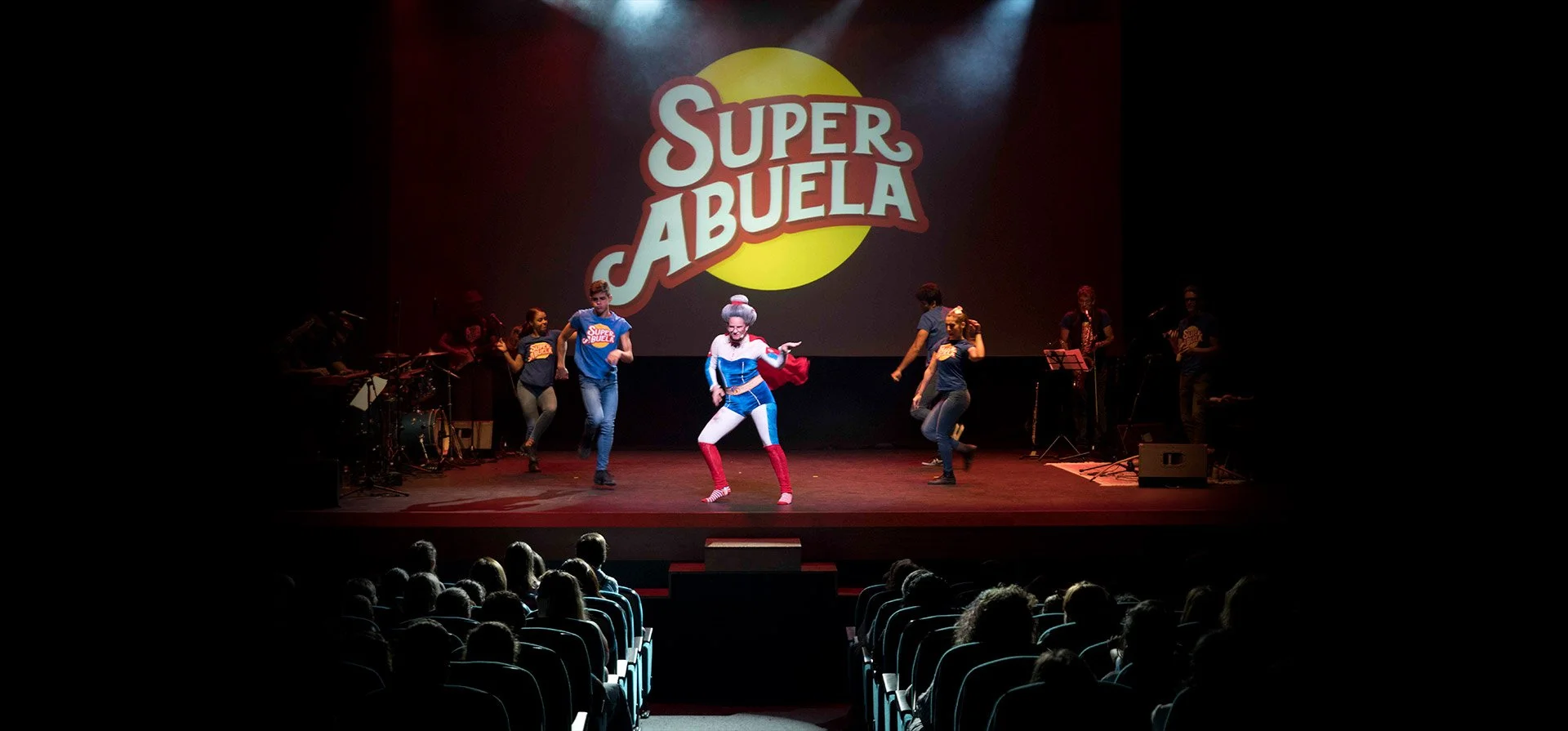Theatrical stage show with costumed characters and live band, with large sign reading 'Super Grandma' on a dark background.