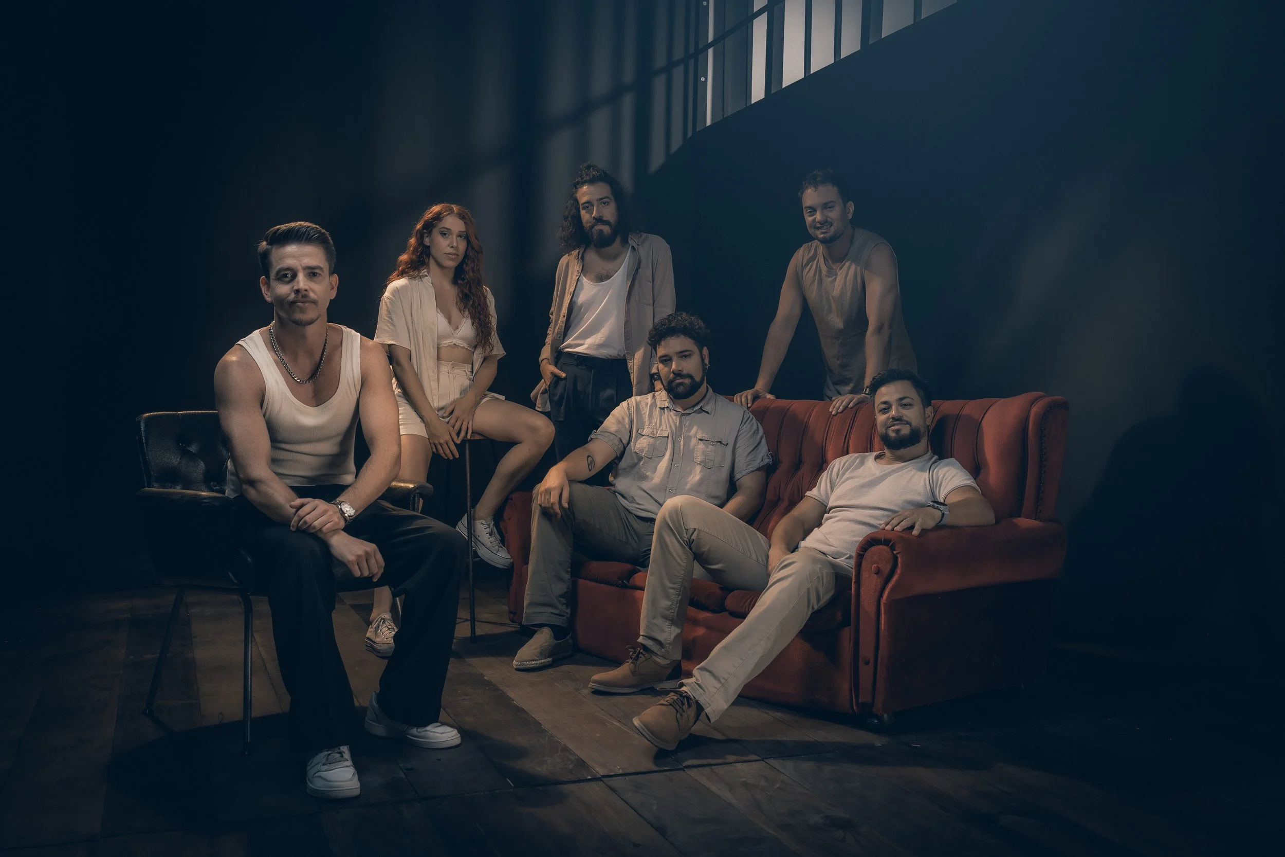 A group of seven people posing in a dark background with light coming through a window, some seated in armchairs and others standing.