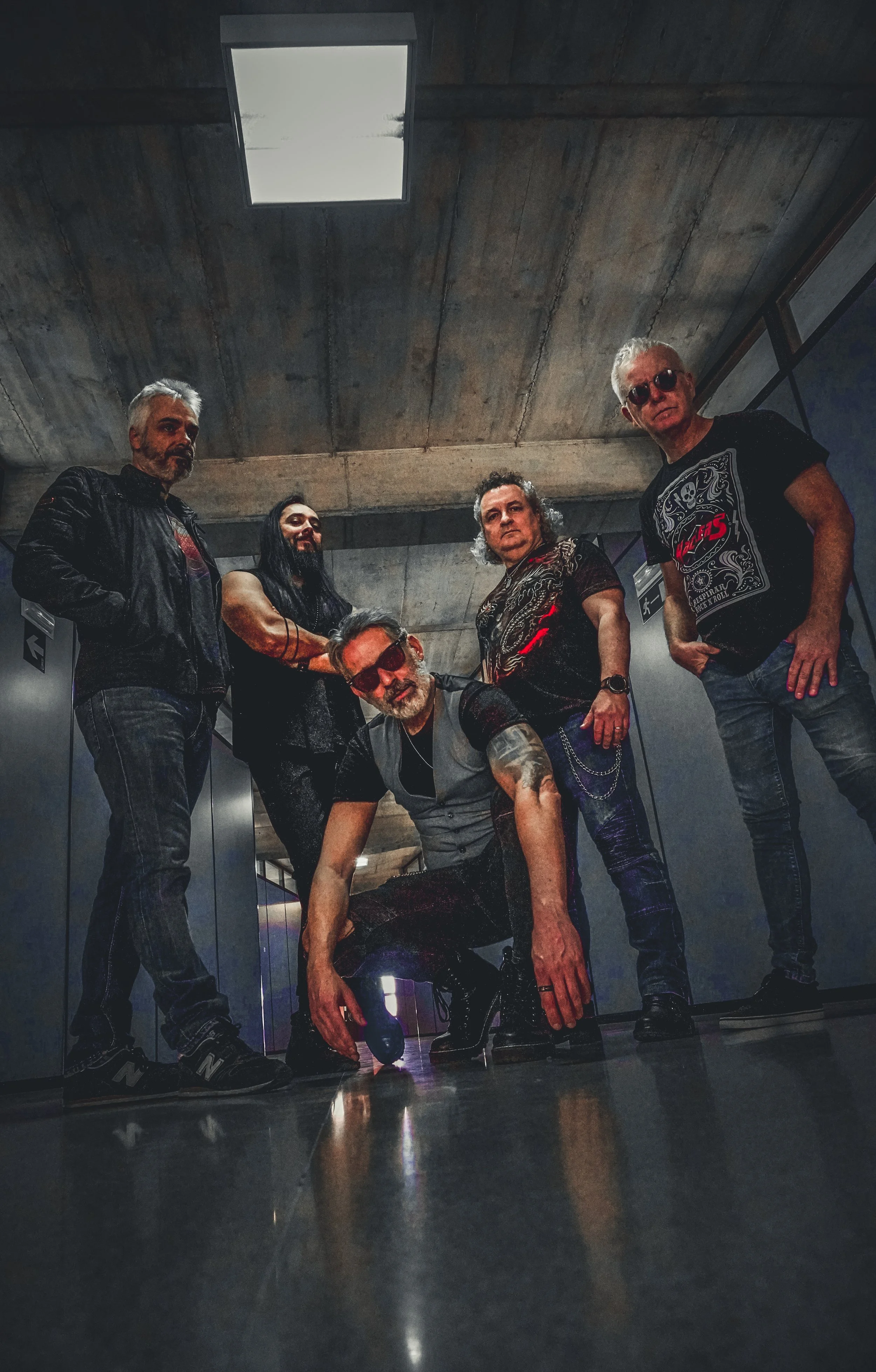 Grupo de cinco personas en un pasillo con techo de concreto, vestidas con ropa casual y estilo rock, posando para la foto.