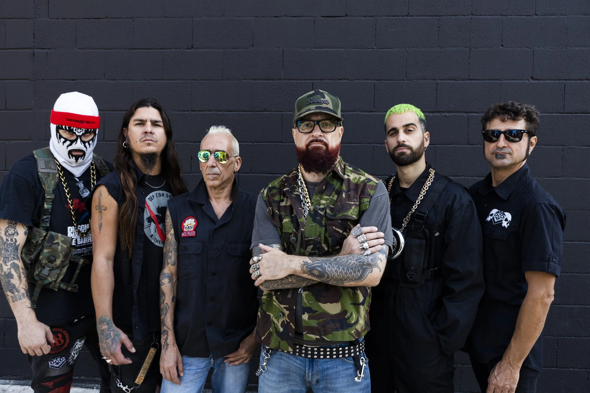 Grupo de seis hombres con estilo urbano, algunos con tatuajes y accesorios llamativos, posando frente a una pared negra.