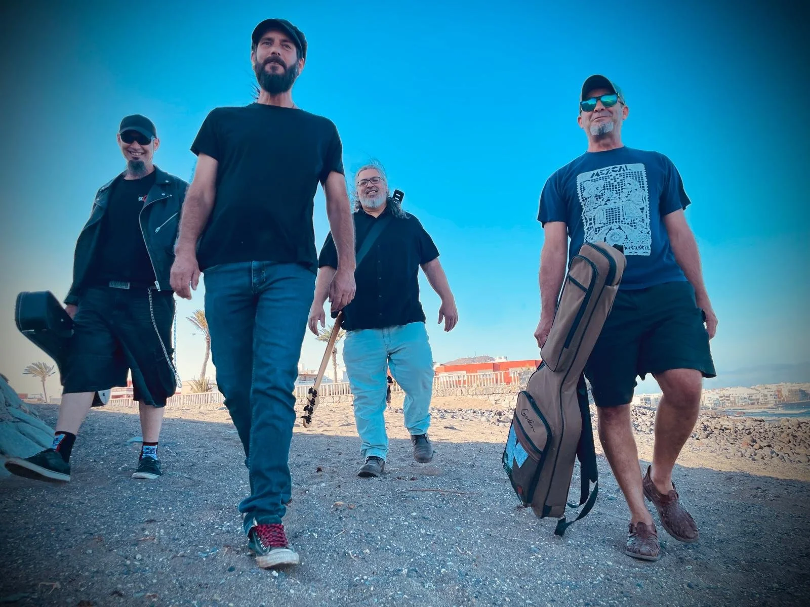 Grupo de cinco hombres caminando por la playa con equipos de guitarra y maletines, disfrutando del día de playa