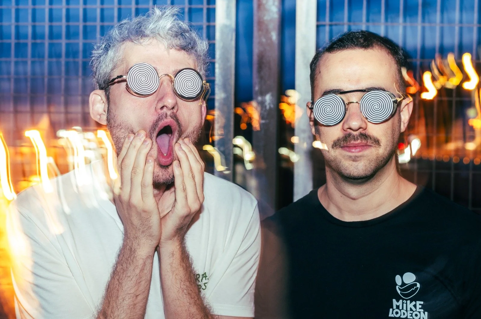 Dos hombres con gafas con espirales en los lentes, uno con expresión de sorpresa y el otro con expresión neutral, en un fondo nocturno con luces borrosas.