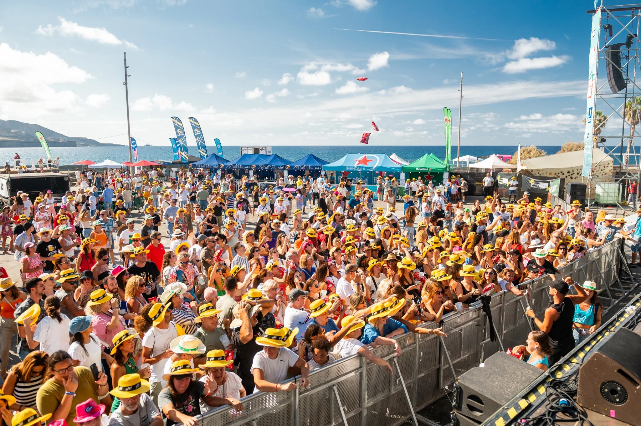 Multitud de personas en un festival en la playa usando sombreros amarillos, con carpas, banderas y vista al mar bajo cielo soleado.
