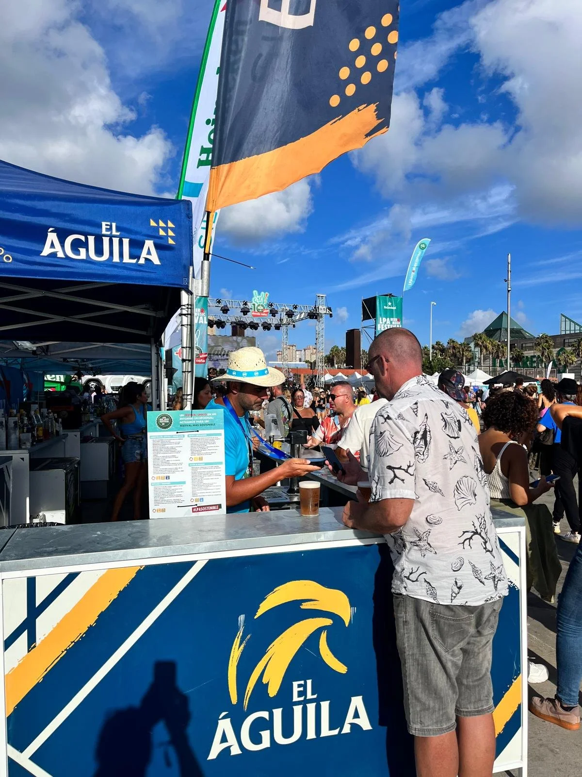 Hombre en camiseta de camiseta con temática marina comprando en un puesto de feria al aire libre. El puesto tiene un cartel con el logotipo de 'El Águila' y un banner en la parte superior con el mismo logo y un diseño de águila en amarillo y azul. Hay muchas personas en el fondo disfrutando del evento y un cielo despejado con algunas nubes.