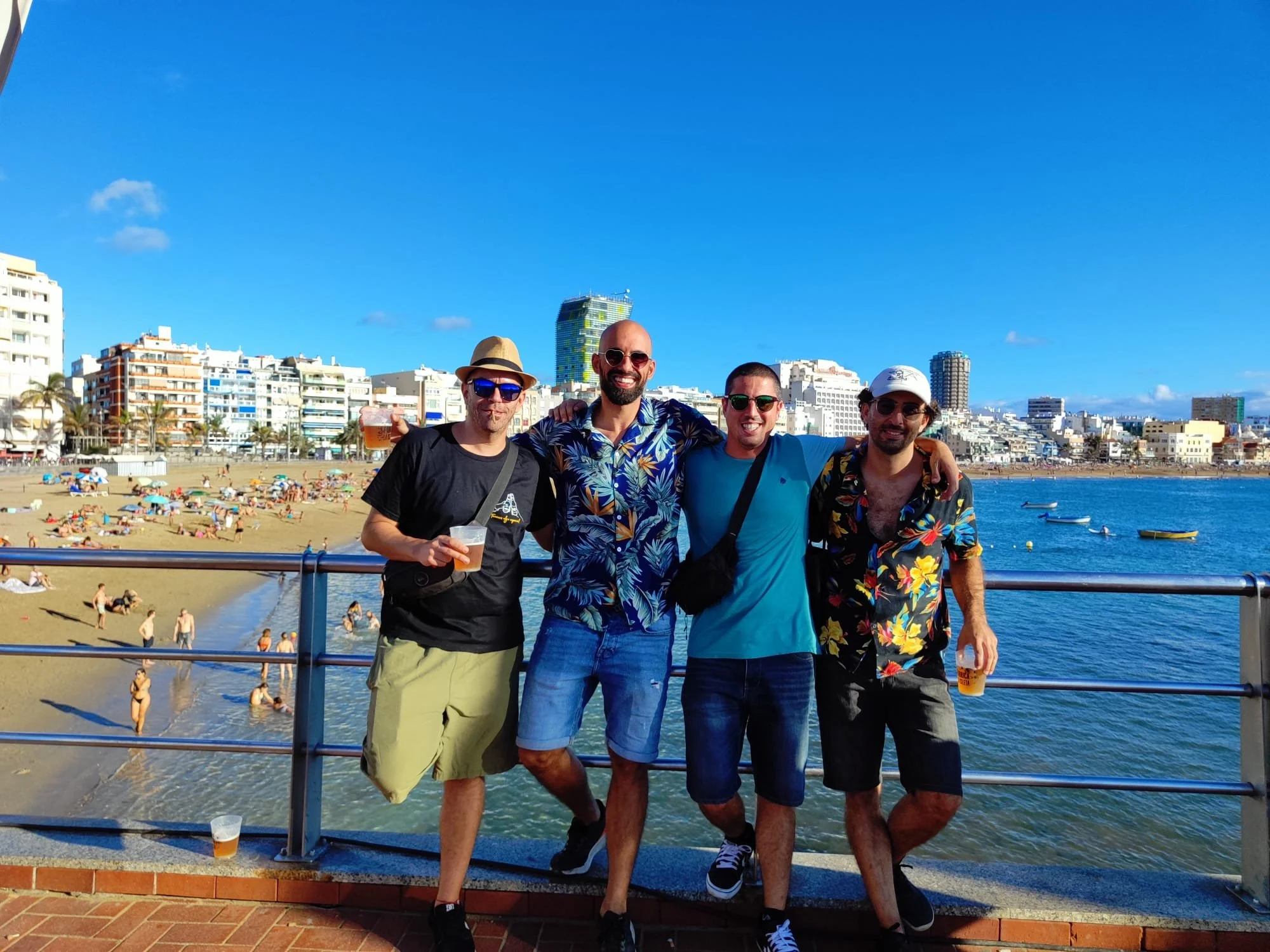 Four friends standing by the sea on a terrace overlooking the beach with urban buildings in the background, enjoying a sunny afternoon with drinks in hand.