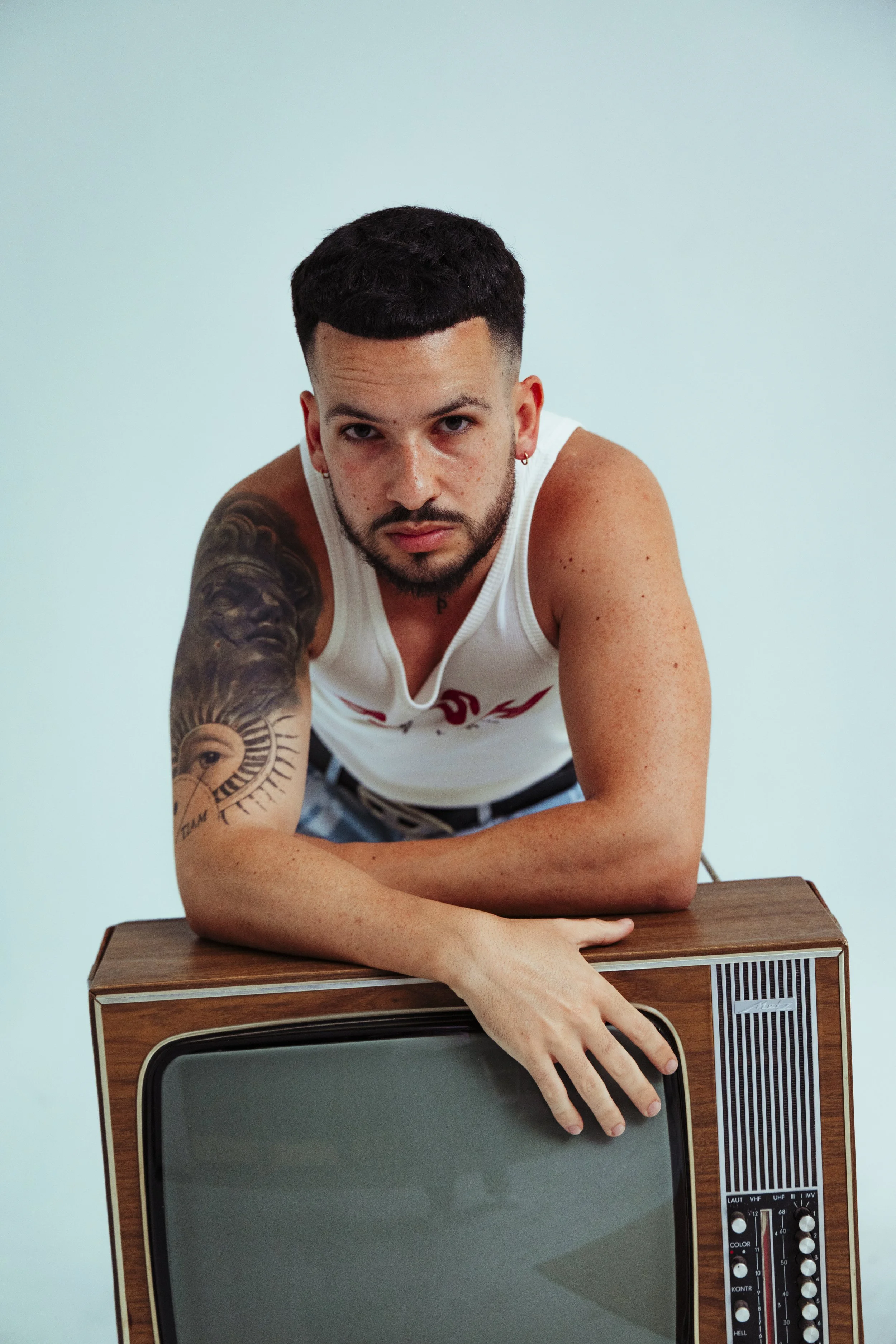 Joven con tatuajes, barba y cabello corto, apoyando sus brazos sobre un televisor de madera, usando una camiseta blanca y pantalones cortos.