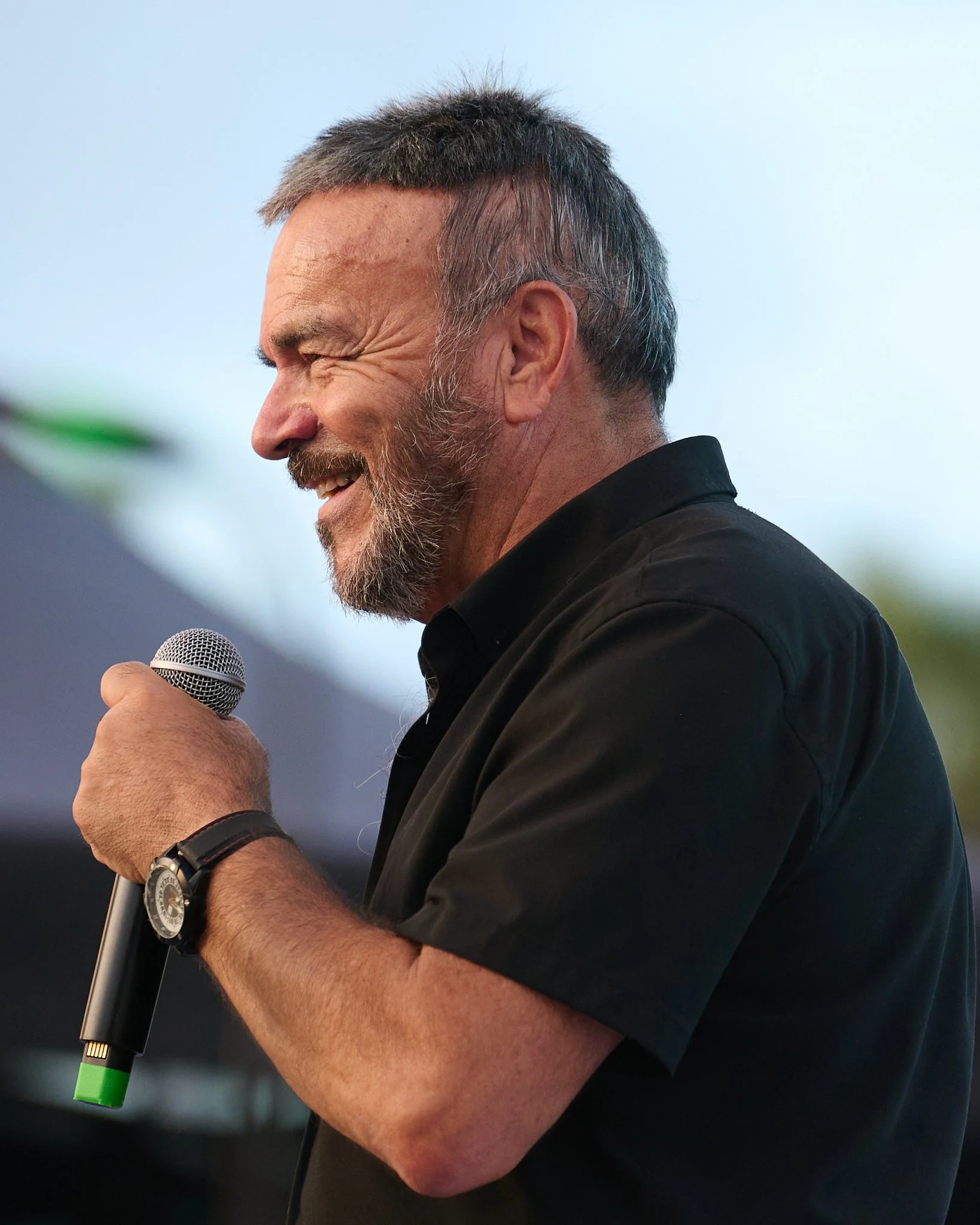 Bearded man holding a microphone and smiling, dressed in a black T-shirt, during an outdoor conversation or presentation.