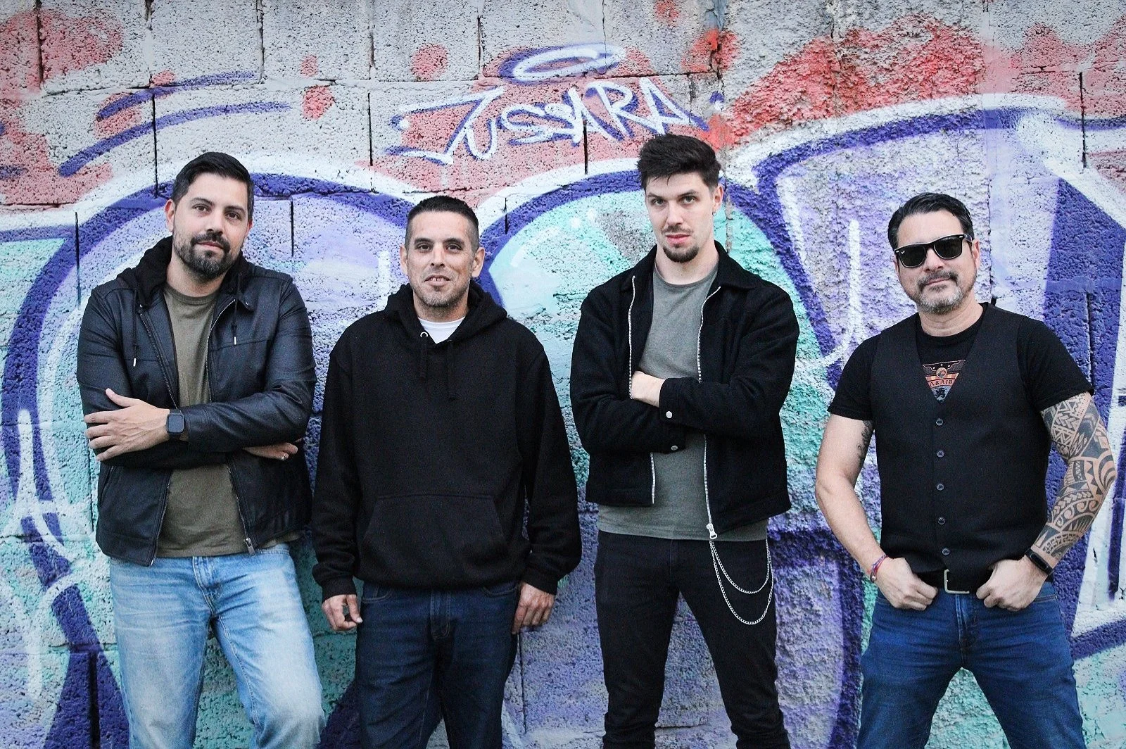 Cuatro hombres en una pared de graffiti, uno con chaqueta de cuero, otro con sudadera, otro con chaqueta y cadenas, el cuarto con gafas de sol y tatuajes.