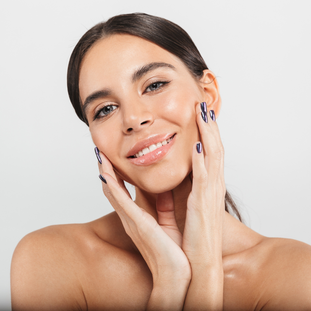 A woman with long dark hair, smiling softly, touching her face with both hands, wearing dark metallic nail polish, against a plain light background.