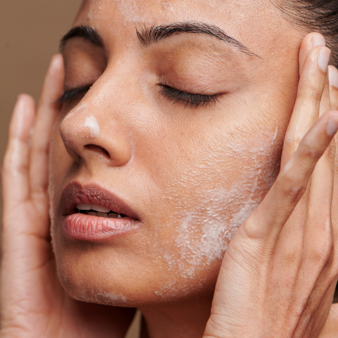 Close-up of a woman with her eyes closed, applying or removing a skincare product from her cheek, with visible moisture on her face.