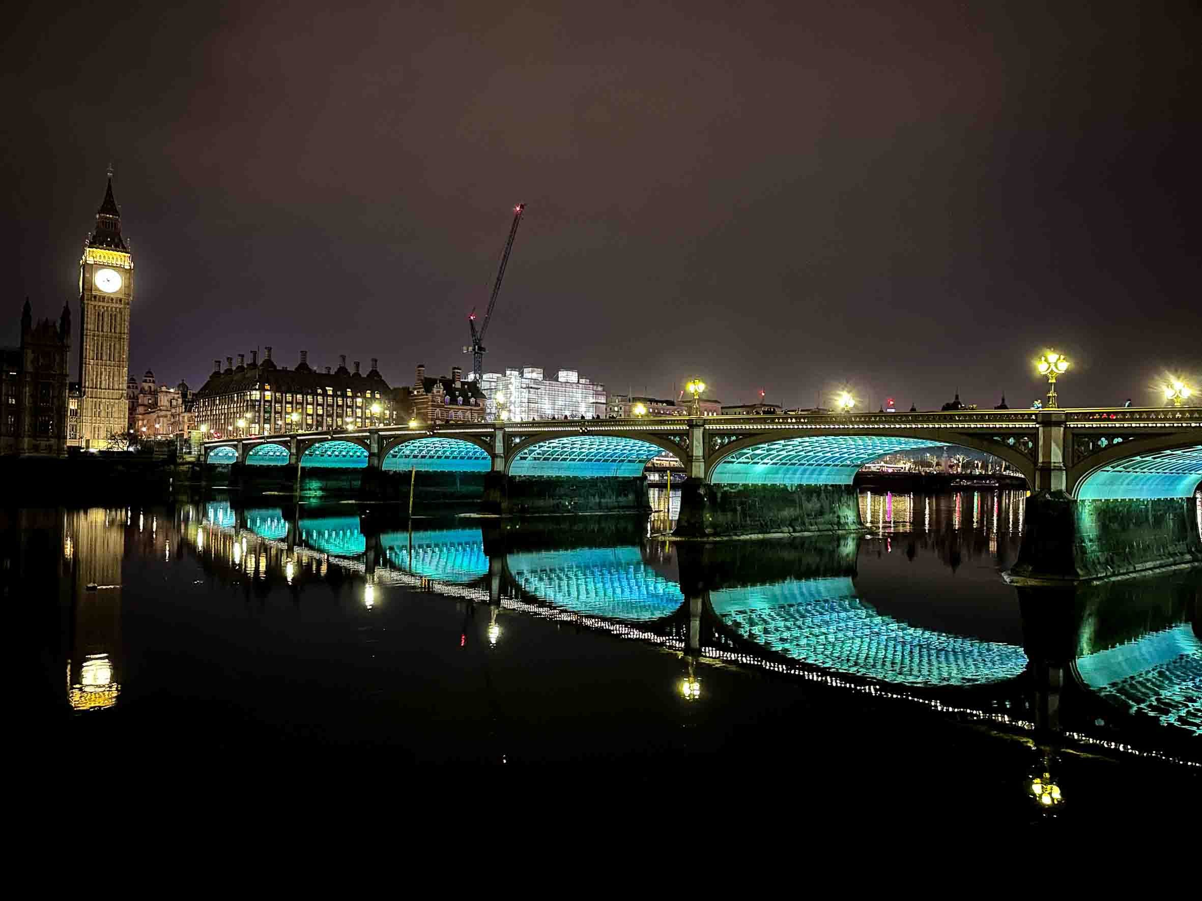 Night view of Westminster Bridge and Big Ben in London, illuminated with city lights and their reflections on the river.