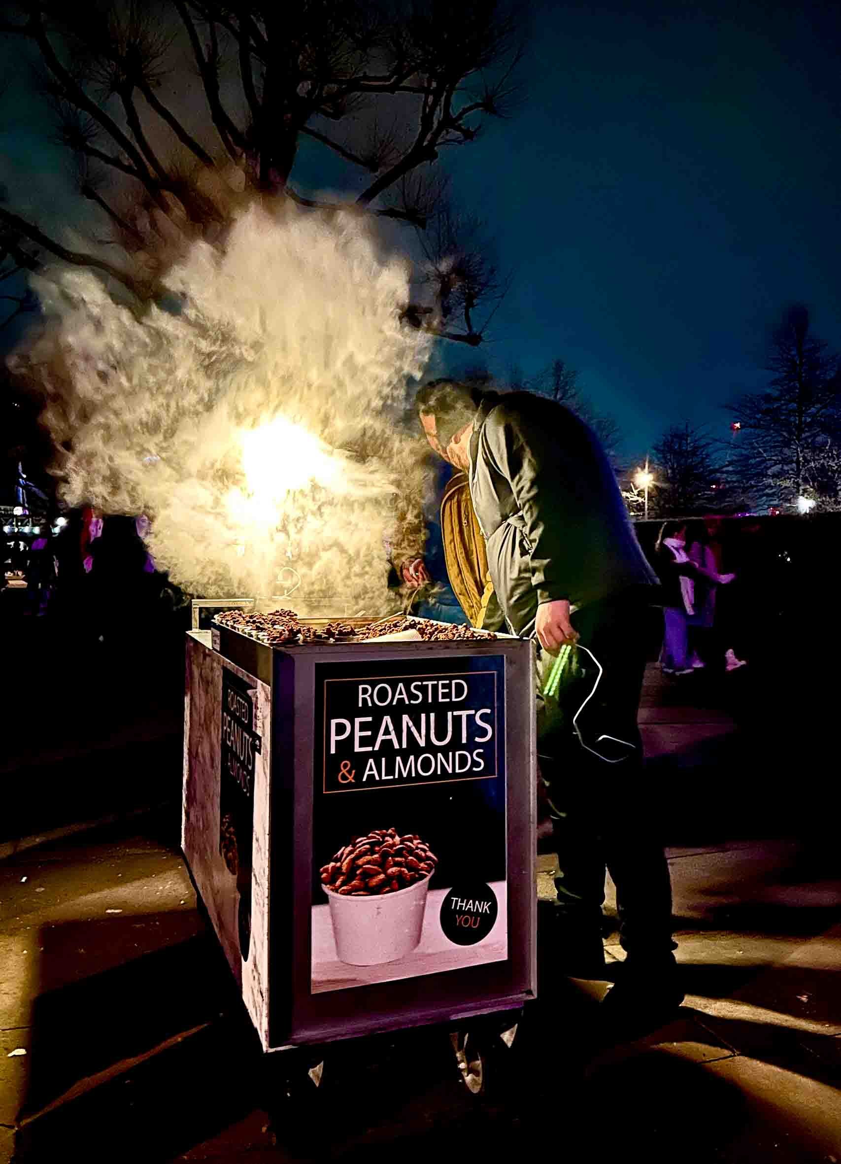 A man in a jacket standing by a cart selling roasted peanuts and almonds at night, with smoke rising from the food and trees in the background.