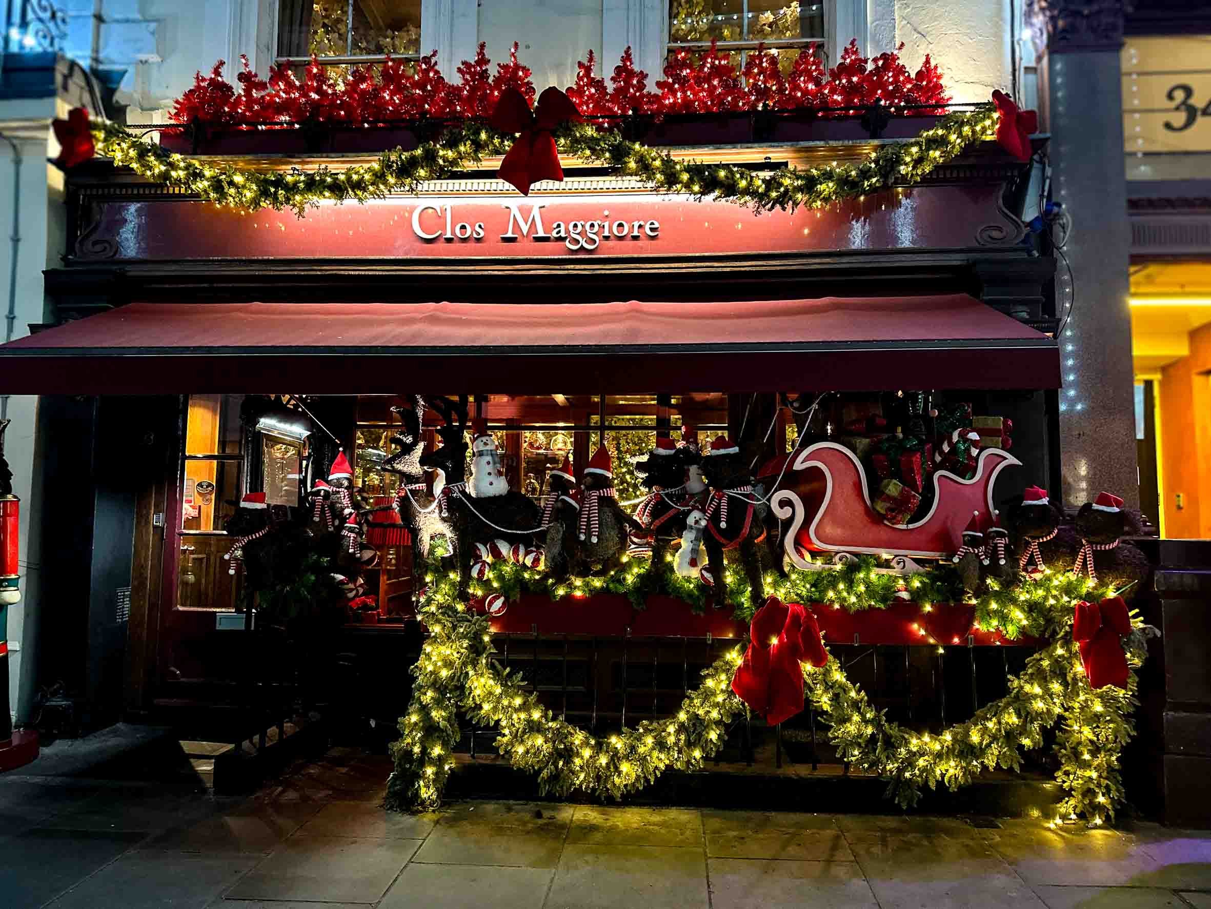 A decorated storefront with Christmas garlands, lights, and holiday figures, including snowmen, reindeer, and a sleigh, with a sign reading 'Clos Maggiore'.