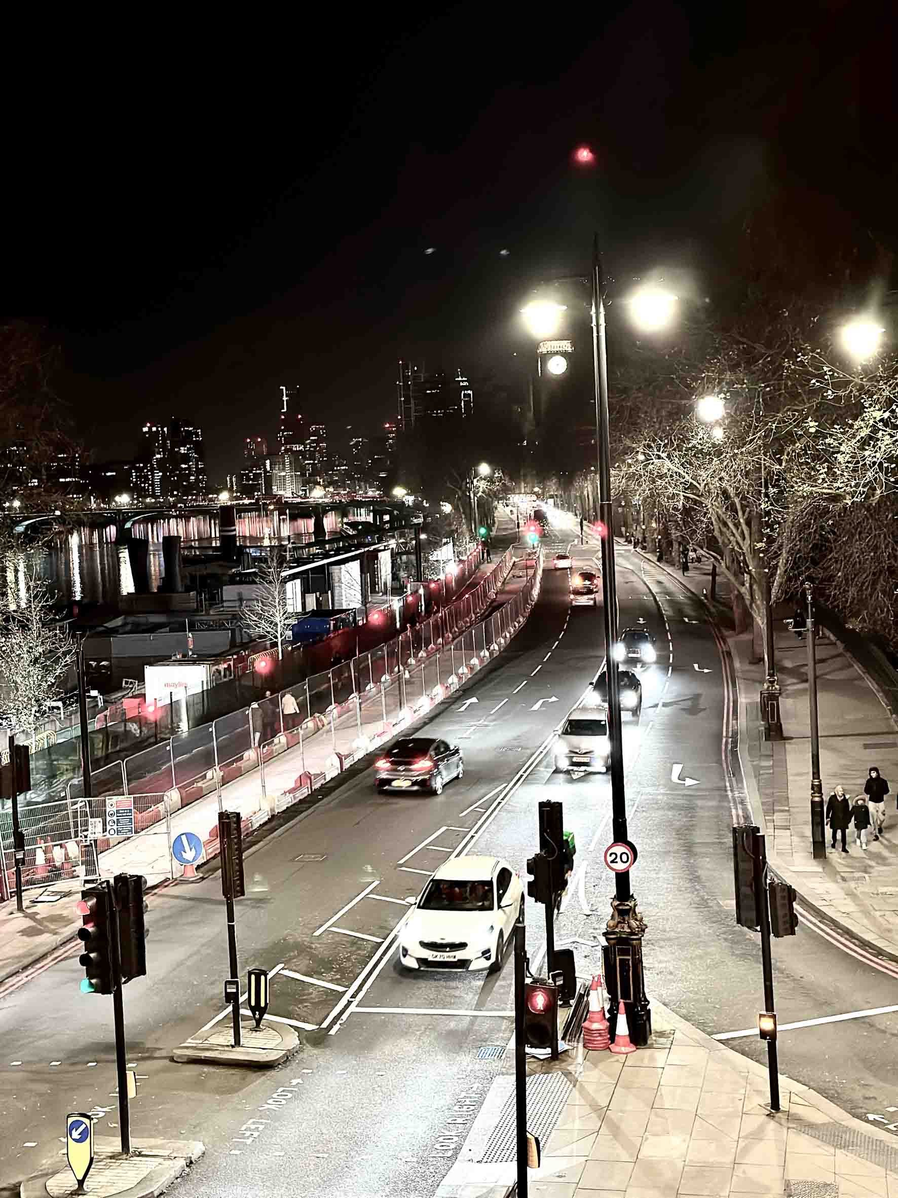Night view of a city street with moving cars, traffic lights, street lamps, and pedestrians, with a skyline in the background.