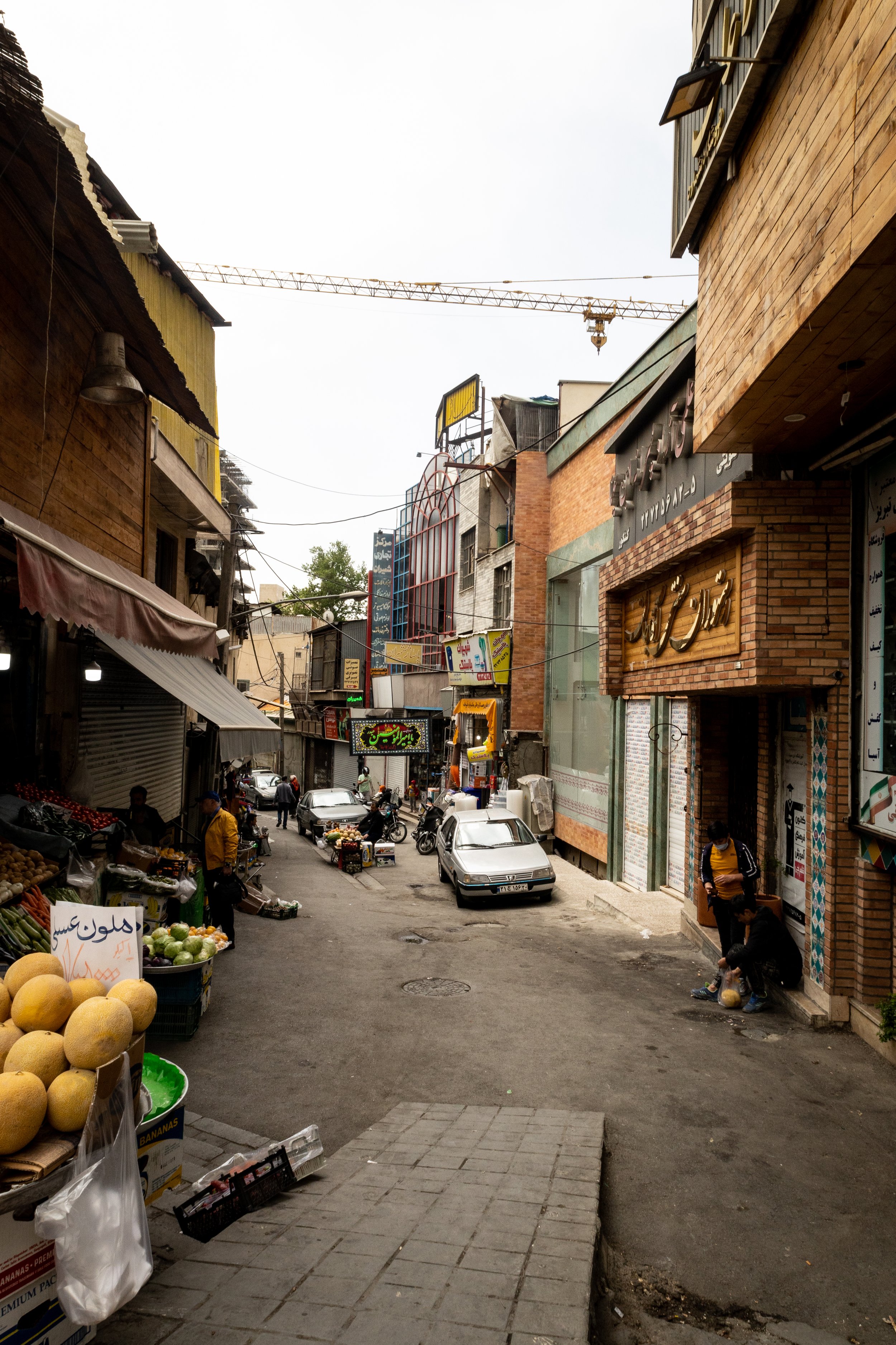 A narrow city street with parked cars and vendors selling fruits and vegetables, with buildings and signage on either side under an overcast sky.