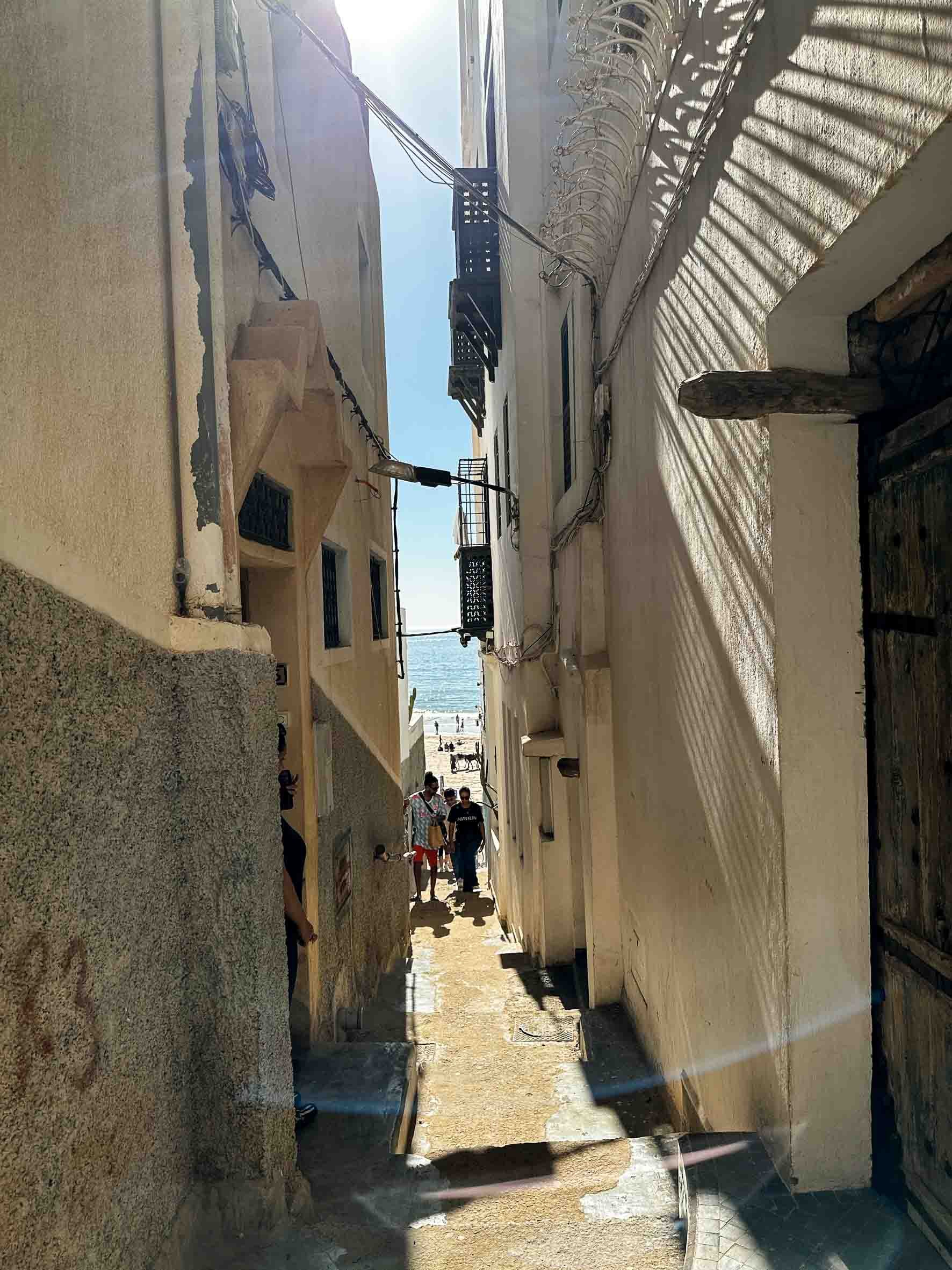 Narrow alleyway leading to the beach with people walking and the ocean in the background.