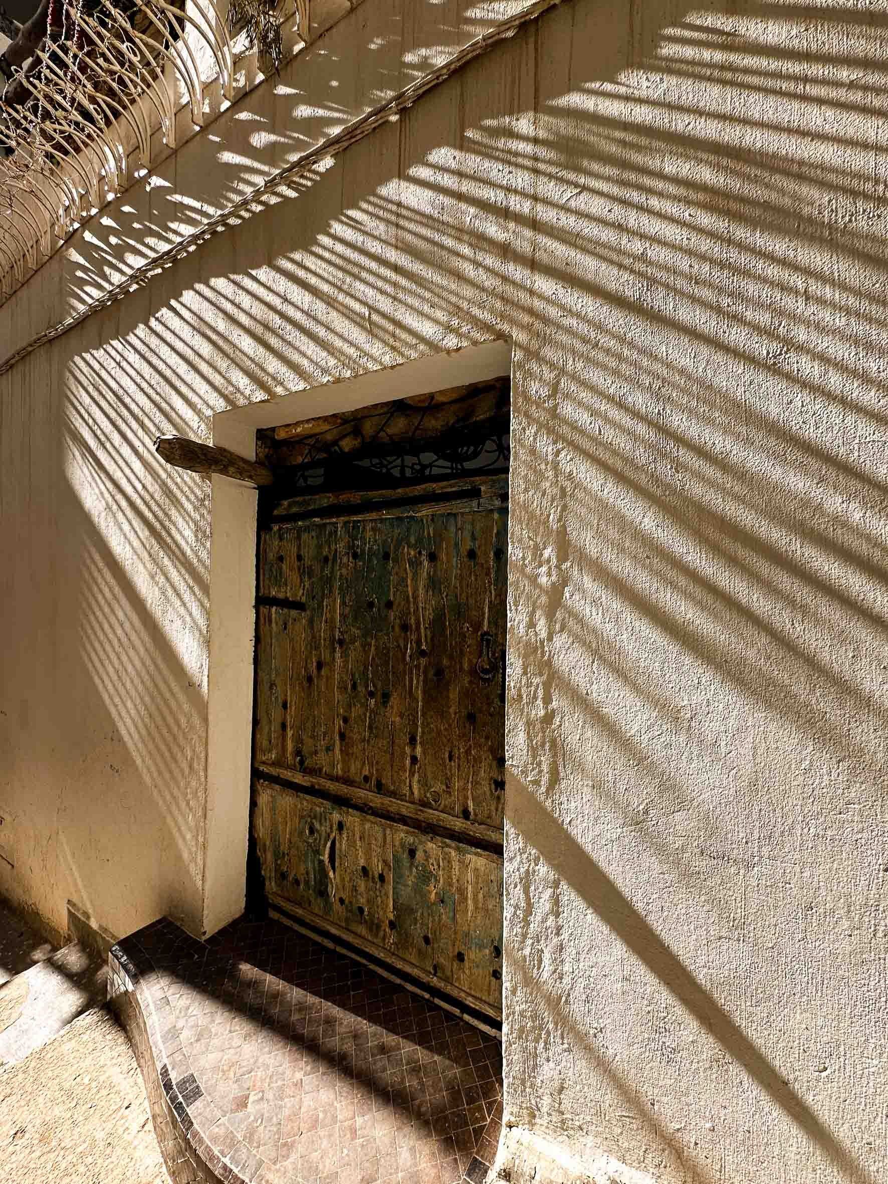 Wooden door with black metal hardware, set in a textured beige wall, with patterned shadow lines from a nearby fence cast on the wall.