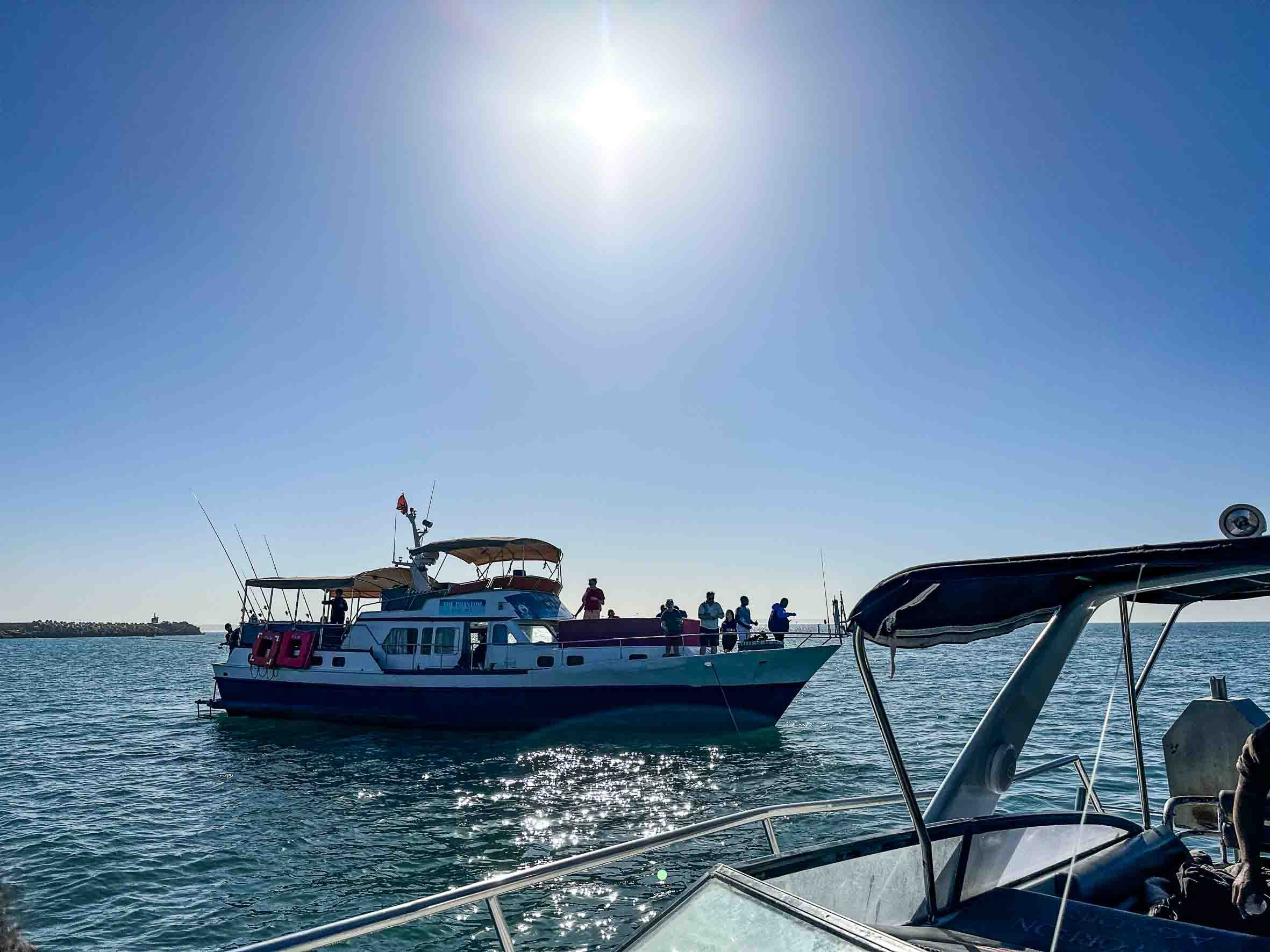 A group of people on a boat enjoying a sunny day on the water, with another boat visible in the foreground and a clear blue sky overhead.