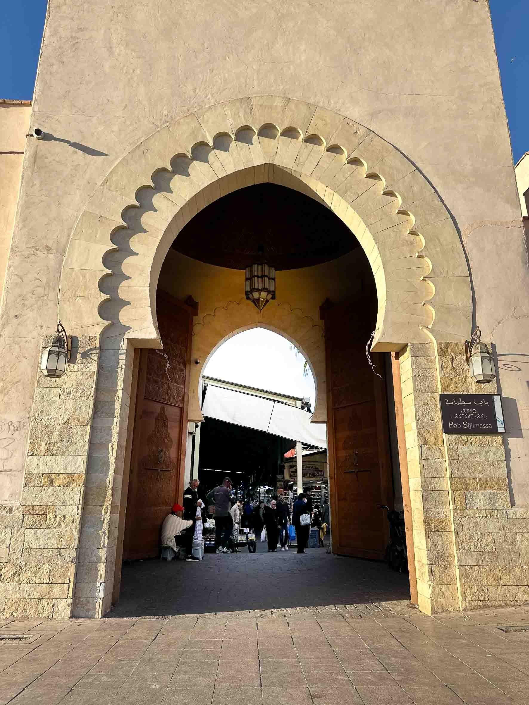 View of Bab Sijilmassa gate at a busy marketplace, with people walking inside and shops visible in the background, featuring traditional Moroccan architecture.