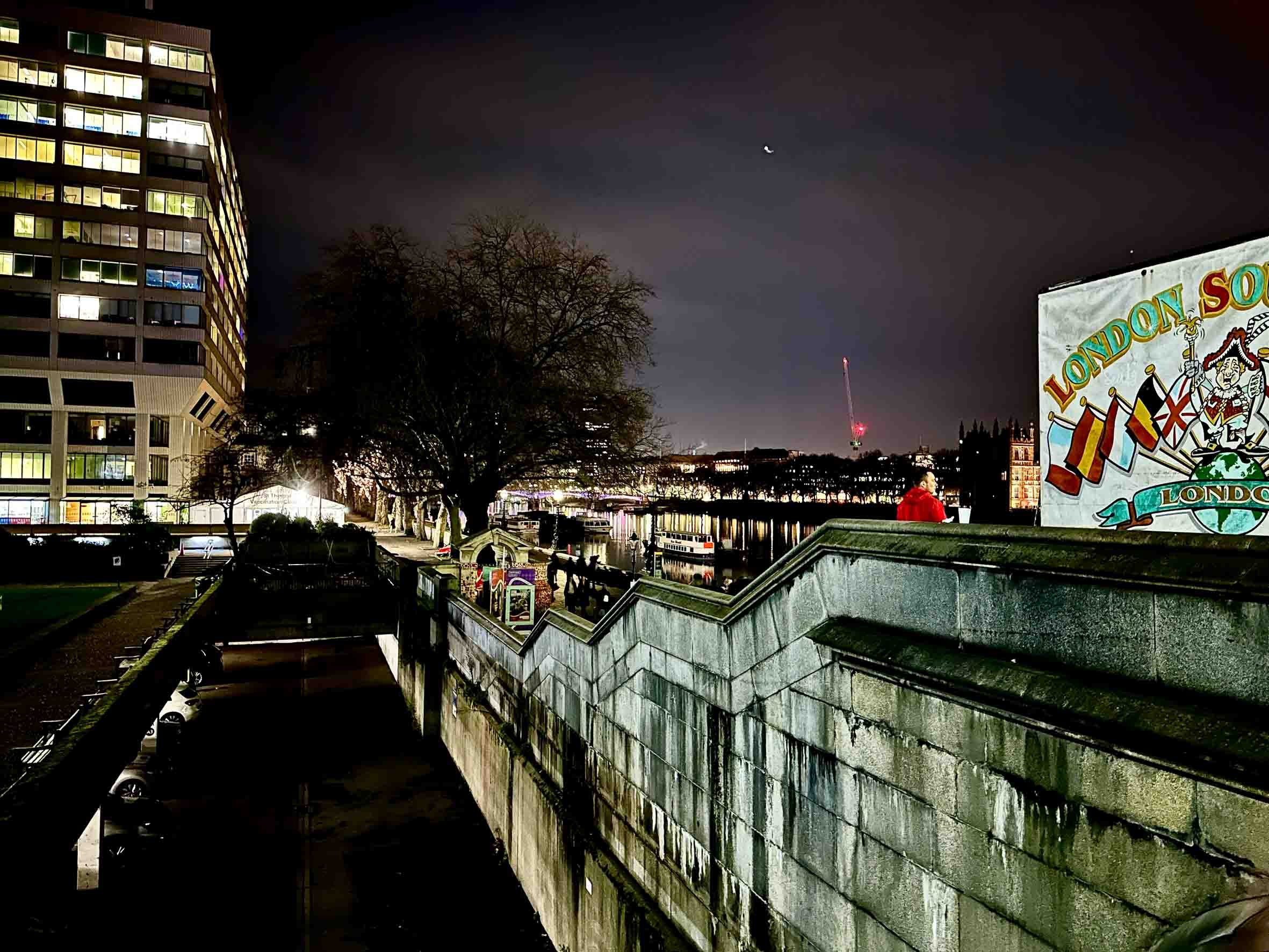 Nighttime view of a waterfront in London with modern and historic buildings, trees, reflections on the water, and a London Soccer banner on the right side.