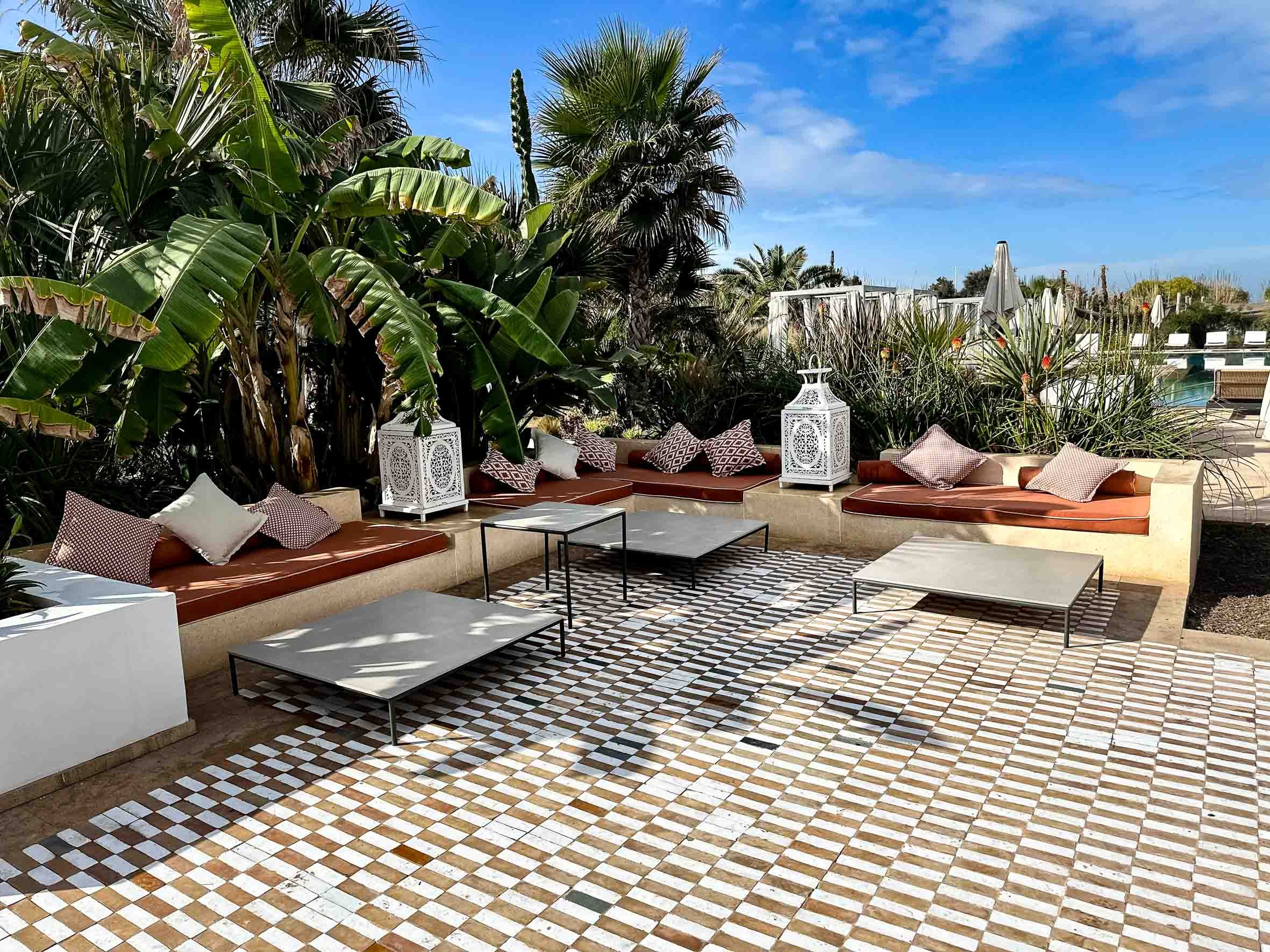 Outdoor patio area with brown cushioned benches, pillows, white lanterns, metal tables, a checkered tile floor, lush green plants, and a swimming pool in the background under a blue sky.