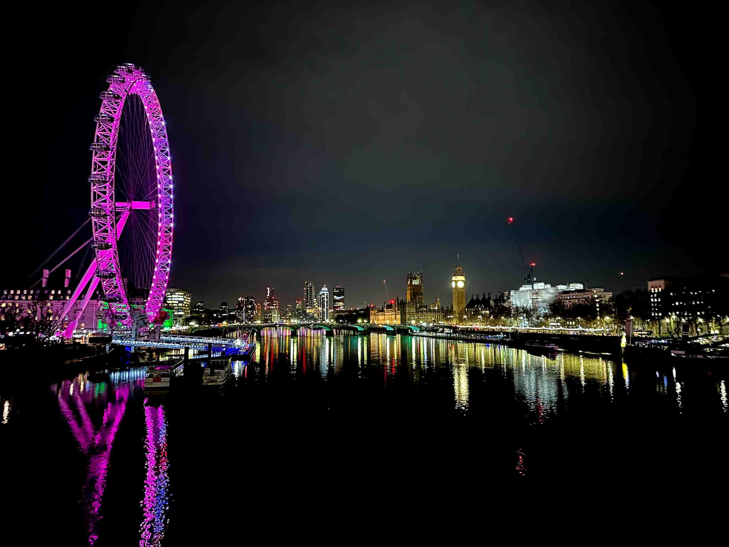 Night view of London skyline featuring the illuminated London Eye Ferris wheel in pink and purple lights, the Big Ben clock tower, and the Houses of Parliament reflected in the River Thames.